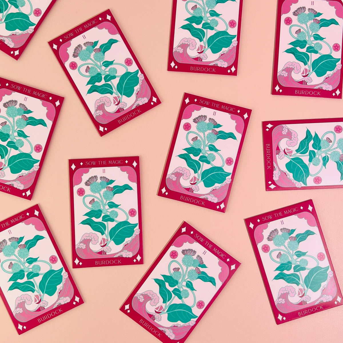 Several Sow the Magic Burdock Tarot Garden Seed Packets are scattered on a pink surface, featuring tarot-style cards with green burdock leaves, purple flowers, seeds, and a decorative pink border.