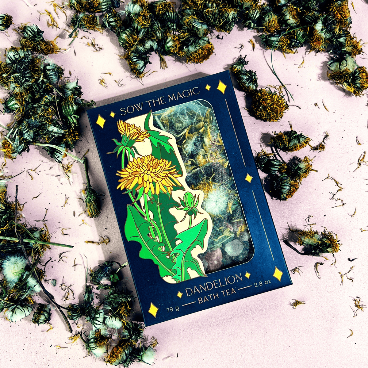 A Sow the Magic Dandelion Tarot Botanical Bath Tea Box, featuring an illustrated dandelion on the front and scattered dried dandelion flowers on a pink surface, highlights detoxifying properties for a rejuvenating bath soak.