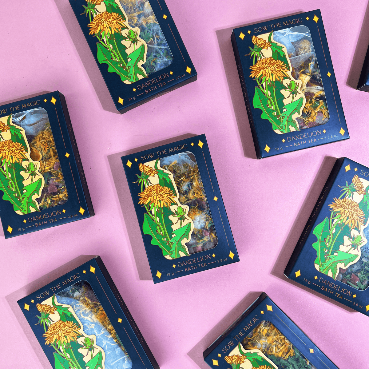 Boxes of Sow the Magic’s Dandelion Tarot Botanical Bath Tea are arranged on a pink surface. The dark blue boxes feature yellow dandelions and highlight the detoxifying, soothing benefits of a botanical tea soak.