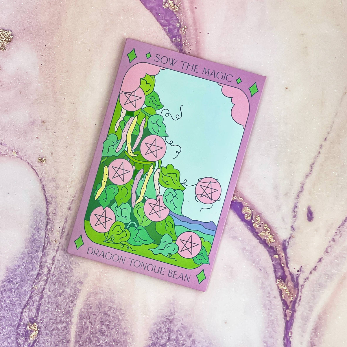 The Sow the Magic Dragon Tongue Bush Bean Tarot Garden Seed Packet features heirloom seeds and a vibrant card with bean plants and pink pentacles, inspired by the Seven of Pentacles, set on a pastel background with purple and gold marbling.