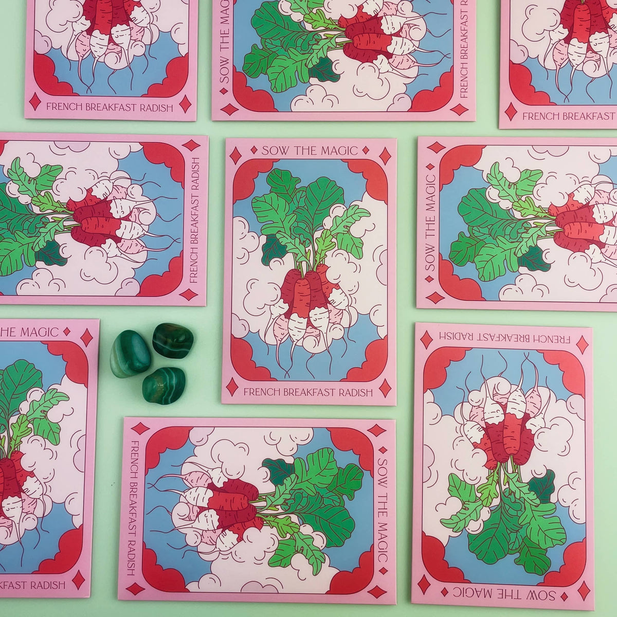 A flat lay of the Sow the Magic French Breakfast Radish Tarot Garden Seed Packet, featuring illustrated bunches of French radish seeds with green leaves, on a mint green background with three green stones.