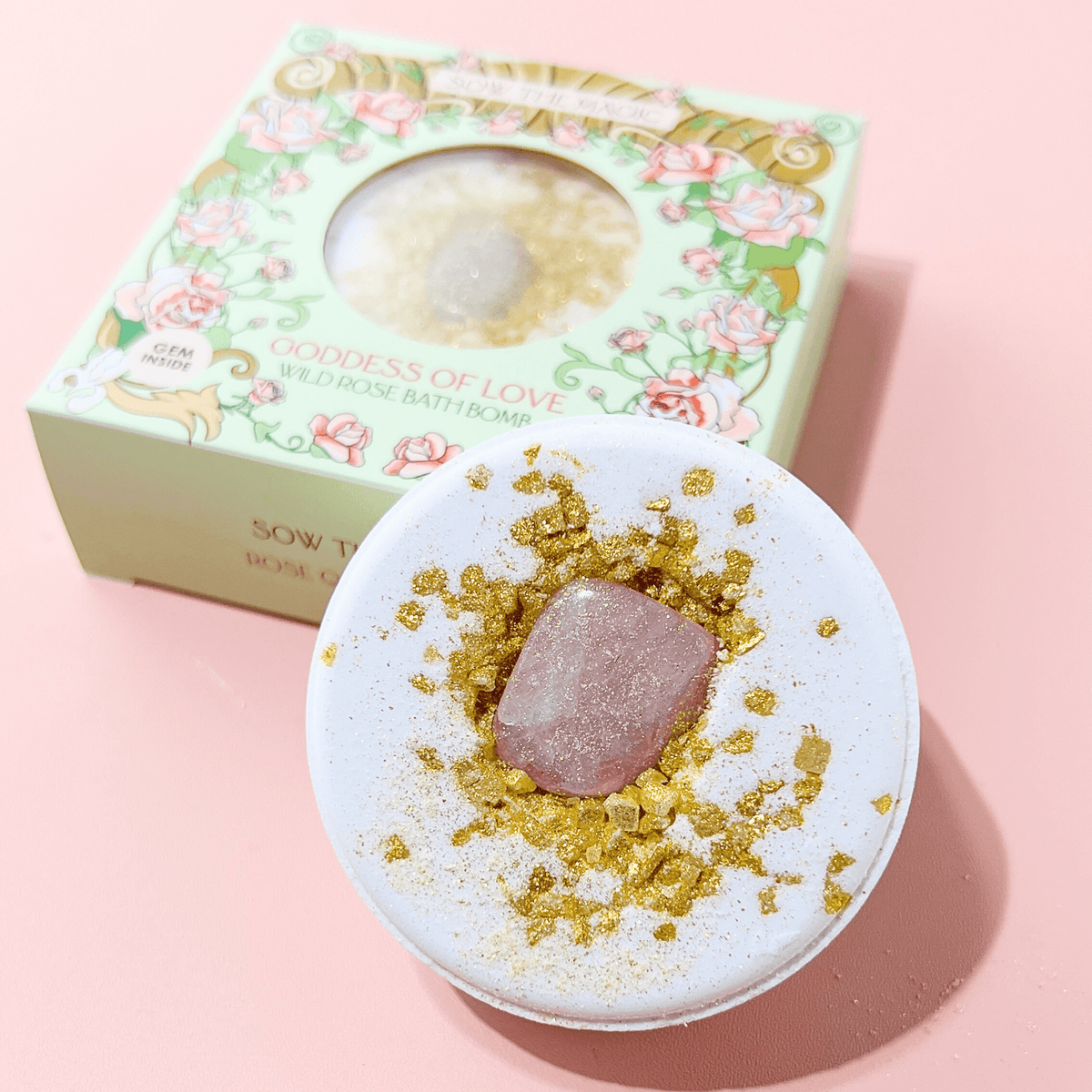 The Sow the Magic Goddess of Love Wild Rose Bath Bomb with Rose Quartz, featuring a floral scent and gold glitter, is displayed before its gorgeous box on a pale pink surface—a perfect self-care treat.