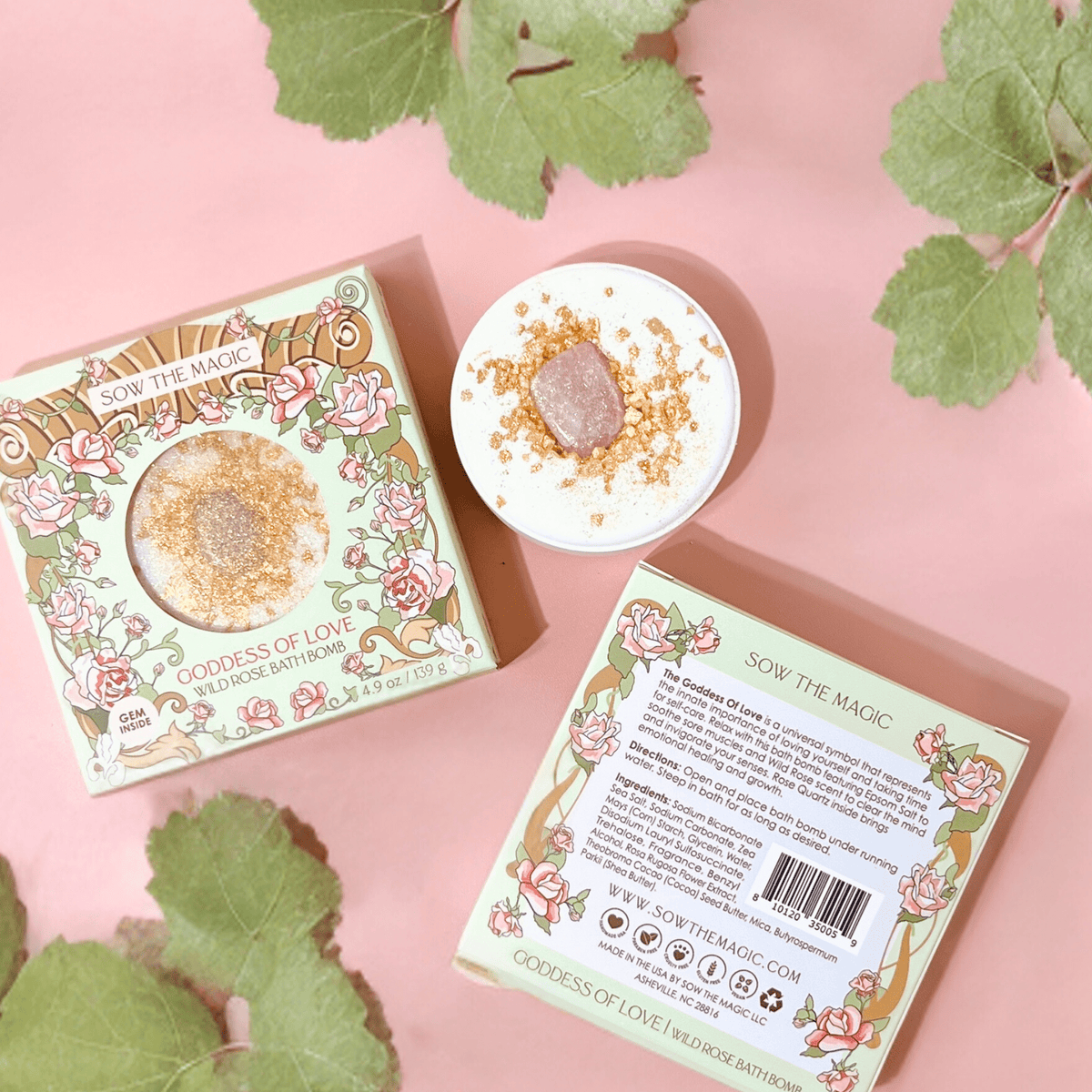 The Sow the Magic Goddess of Love Wild Rose Bath Bomb with Rose Quartz, adorned with gold glitter, rests beside its floral packaging on a pink surface with green leaves, perfect for self-care rituals.