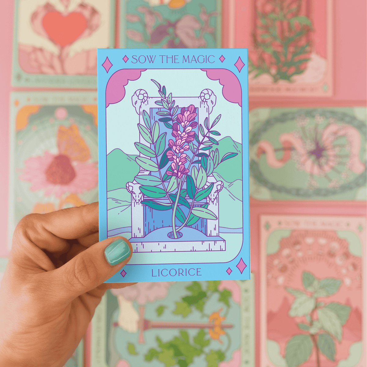 A hand with teal nails holds a vibrant Sow the Magic Licorice Tarot Garden Seed Packet, featuring colorful Glycyrrhiza glabra art. Other Sow the Magic plant seed packets appear blurred in the background.