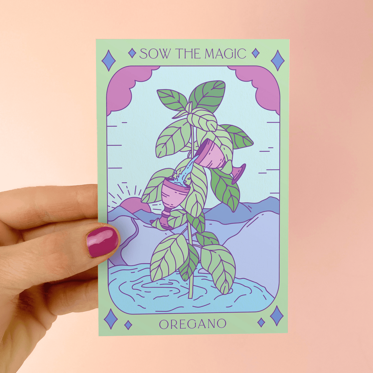 A hand with purple nail polish holds a pastel Sow the Magic Oregano Tarot Garden Seed Packet, featuring illustrated perennial oregano, seeds, watering cans, and a scenic background with hills and a pink sky.
