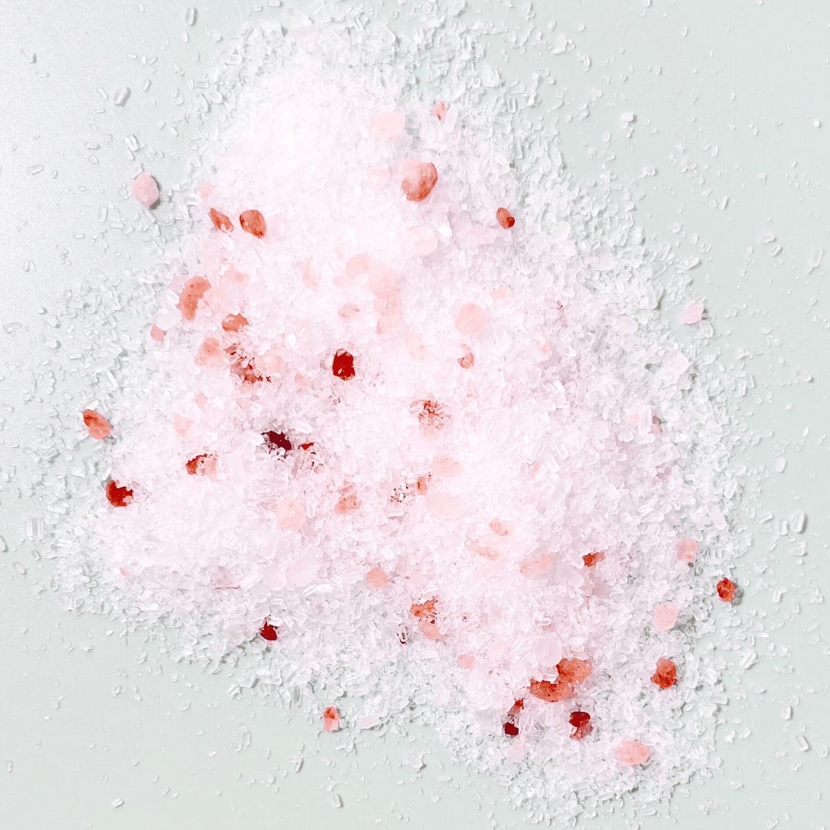 A close-up of The Moon Salt Soak in Lavender by Sow the Magic—coarse white, pink, and red mineral crystals believed to aid intuition and dreams—scattered on a pale light green background.