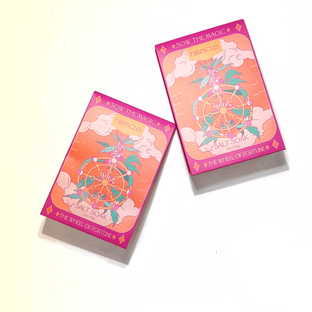 Two rectangular pink and orange boxes labeled Sow the Magic The Wheel of Fortune Salt Soak in Hibiscus, featuring floral and Wheel of Fortune designs, highlight mineral-rich bath salts against a white and soft yellow background.