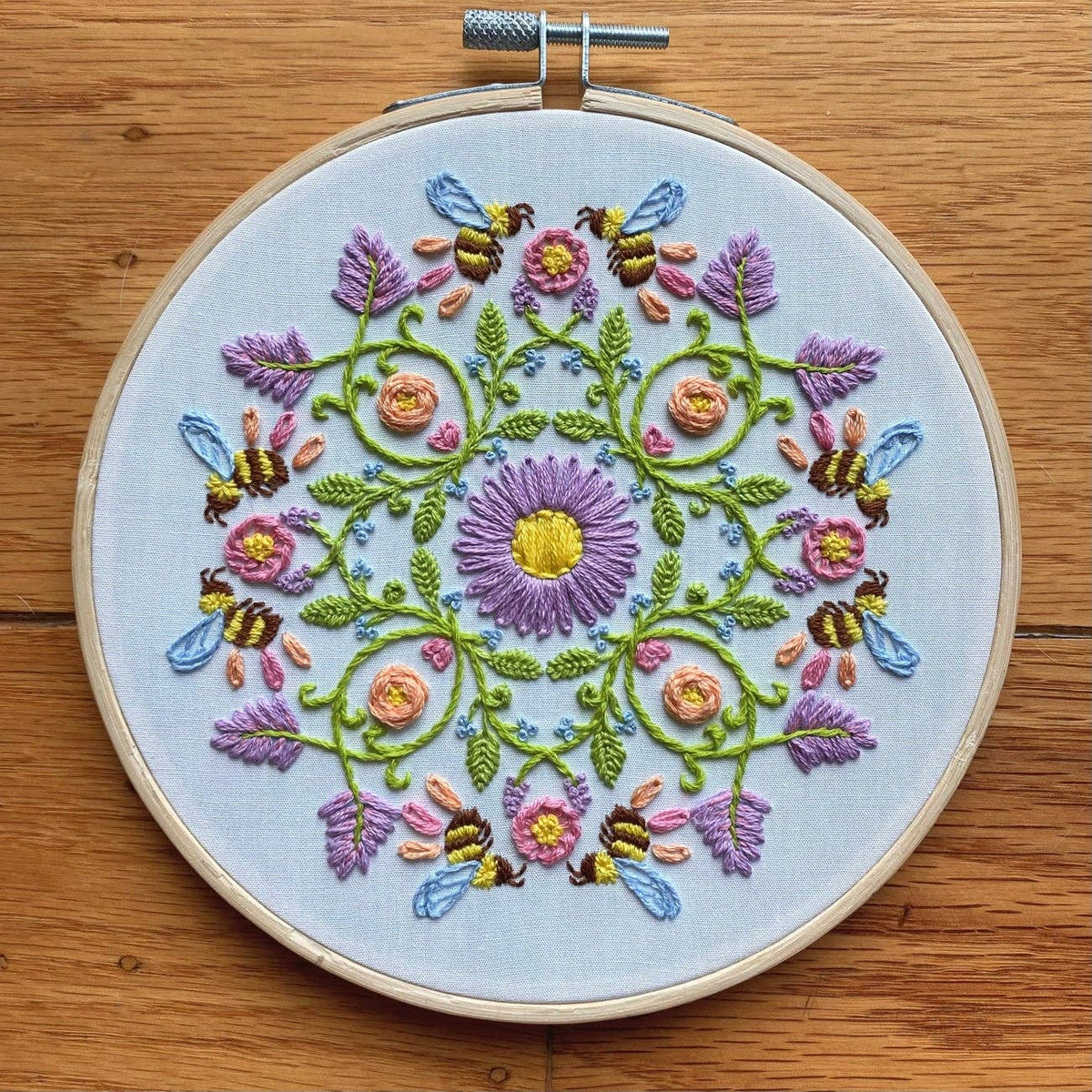 The 6 Bee Mandala DIY Embroidery Kit by Stitches By Tiff features a symmetrical floral design with bees, purple, pink, and yellow flowers on white fabric—ideal for beginners seeking relaxing DIY art projects. Shown on a wooden surface.