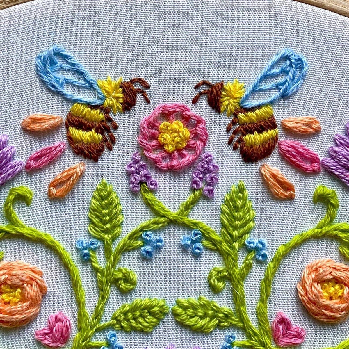 The 6 Bee Mandala DIY Embroidery Kit by Stitches By Tiff features two bees flying to a pink flower, with vibrant blooms and green leaves—ideal for DIY art projects or as inspiration for beginner stitchers.