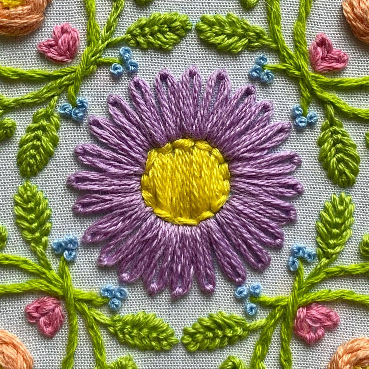 The 6 Bee Mandala DIY Embroidery Kit by Stitches By Tiff features a hand-embroidered flower with purple petals, a yellow center, green leaves, pink flowers, and blue accents on white fabric—perfect for creative art projects.