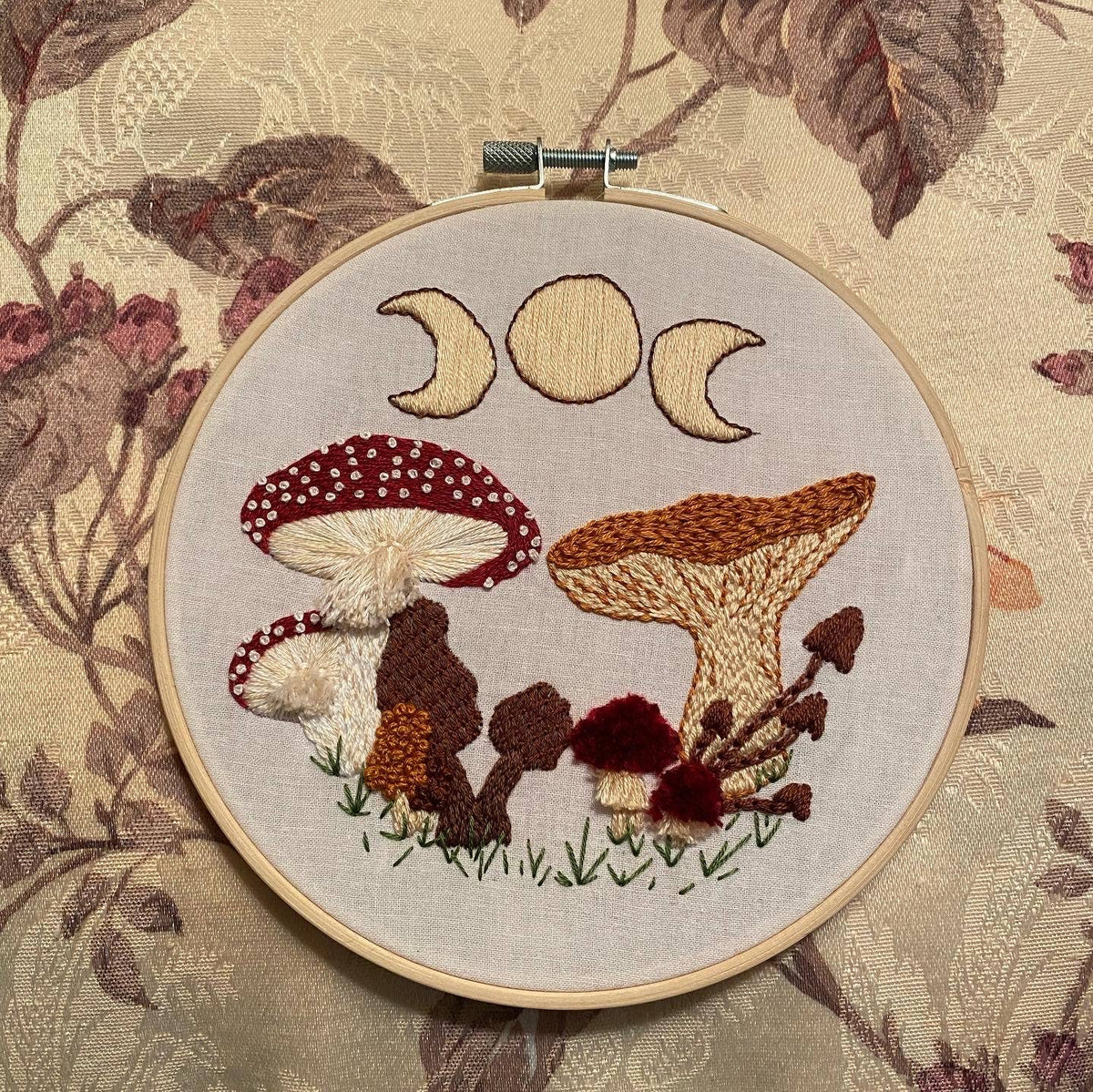 The 6 Beginner Mushie DIY Embroidery Kit by Stitches By Tiff features a mushroom and triple moon design in earthy tones on leafy, floral fabric—ideal for creative home art projects.
