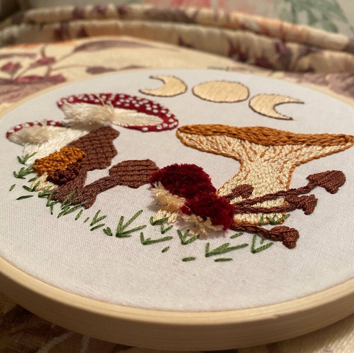 The Stitches By Tiff 6 Beginner Mushie DIY Embroidery Kit features mushrooms, grass, and three crescent moons on white fabric in a wooden hoop—ideal for embroidery beginners or creative DIY art projects.