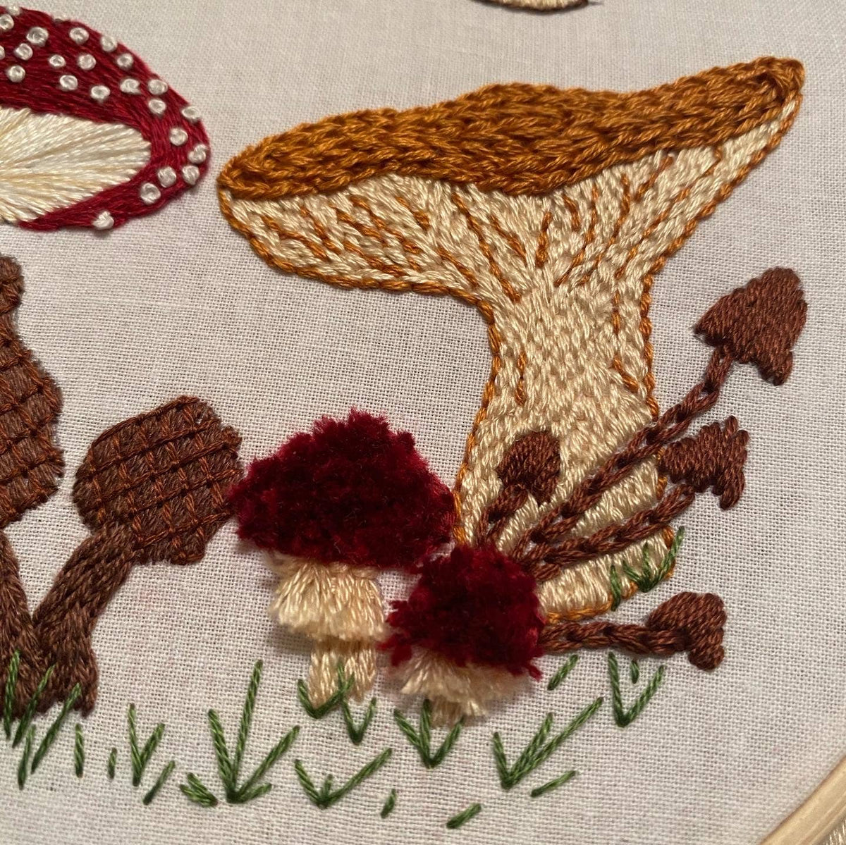 The 6 Beginner Mushie DIY Embroidery Kit from Stitches By Tiff features assorted mushrooms and green grass on fabric—a charming mushroom pattern, perfect for DIY art projects.