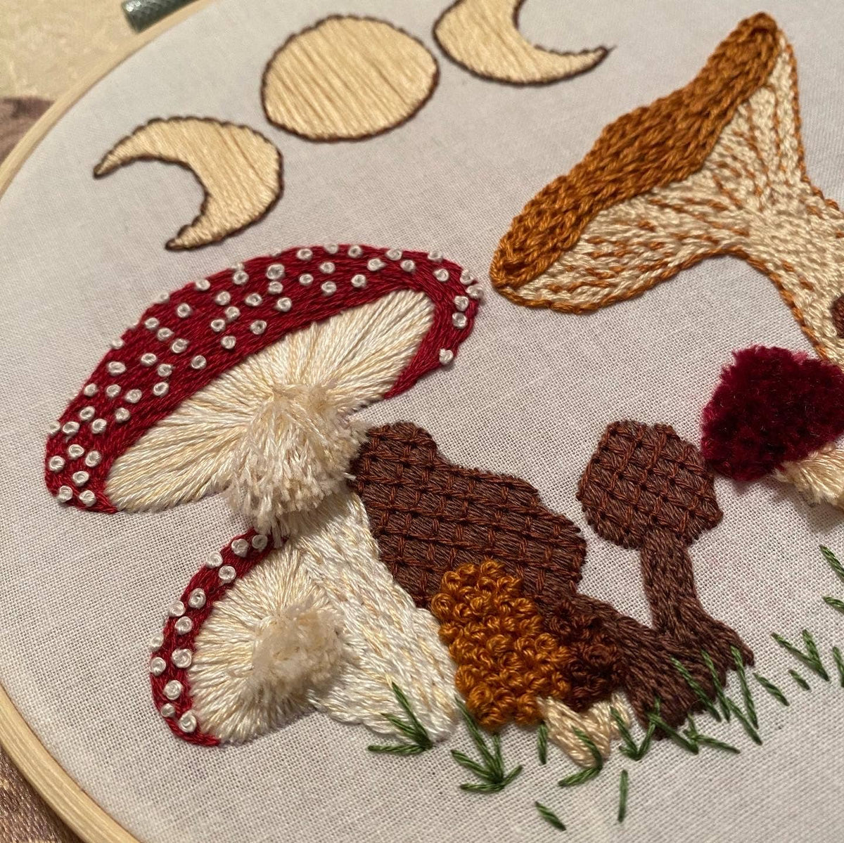 The 6 Beginner Mushie DIY Embroidery Kit by Stitches By Tiff features whimsical red-capped, brown, and yellow mushrooms with grass and crescent moons in beige thread—ideal for creative embroidery projects.