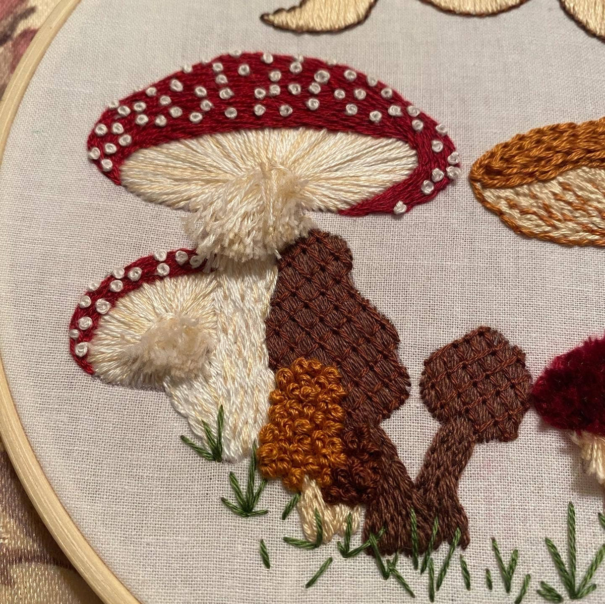 The Stitches By Tiff 6 Beginner Mushie DIY Embroidery Kit features stitched red and white mushrooms, brown fungi, and colorful trees on green grass—ideal inspiration for DIY art projects.