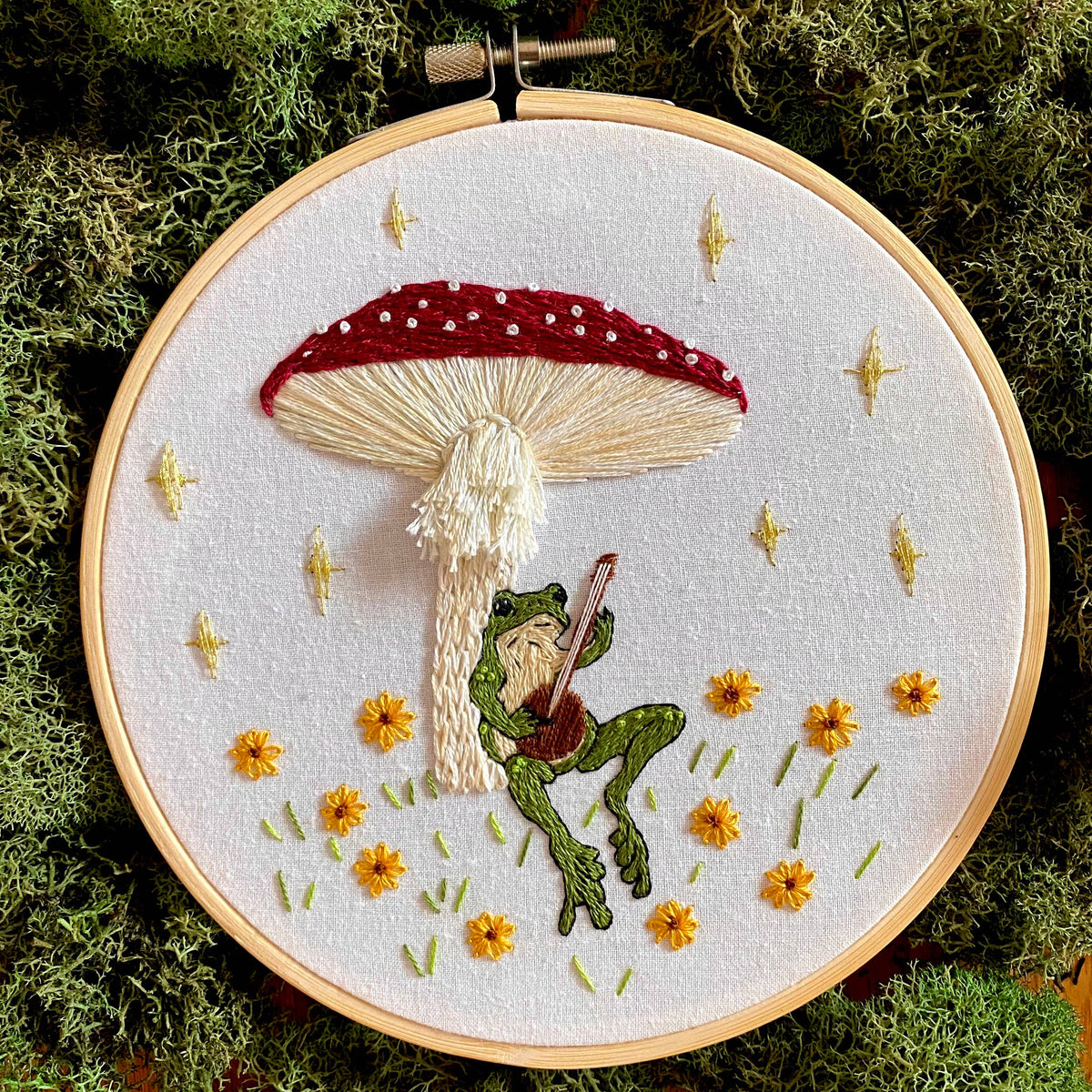 The 6 Beginner Toadstool Serenade Frog & Mushroom DIY Embroidery Kit by Stitches By Tiff features a banjo-playing frog resting on a red-capped mushroom, with yellow flowers and stars—a charming project for fans of whimsical designs!.