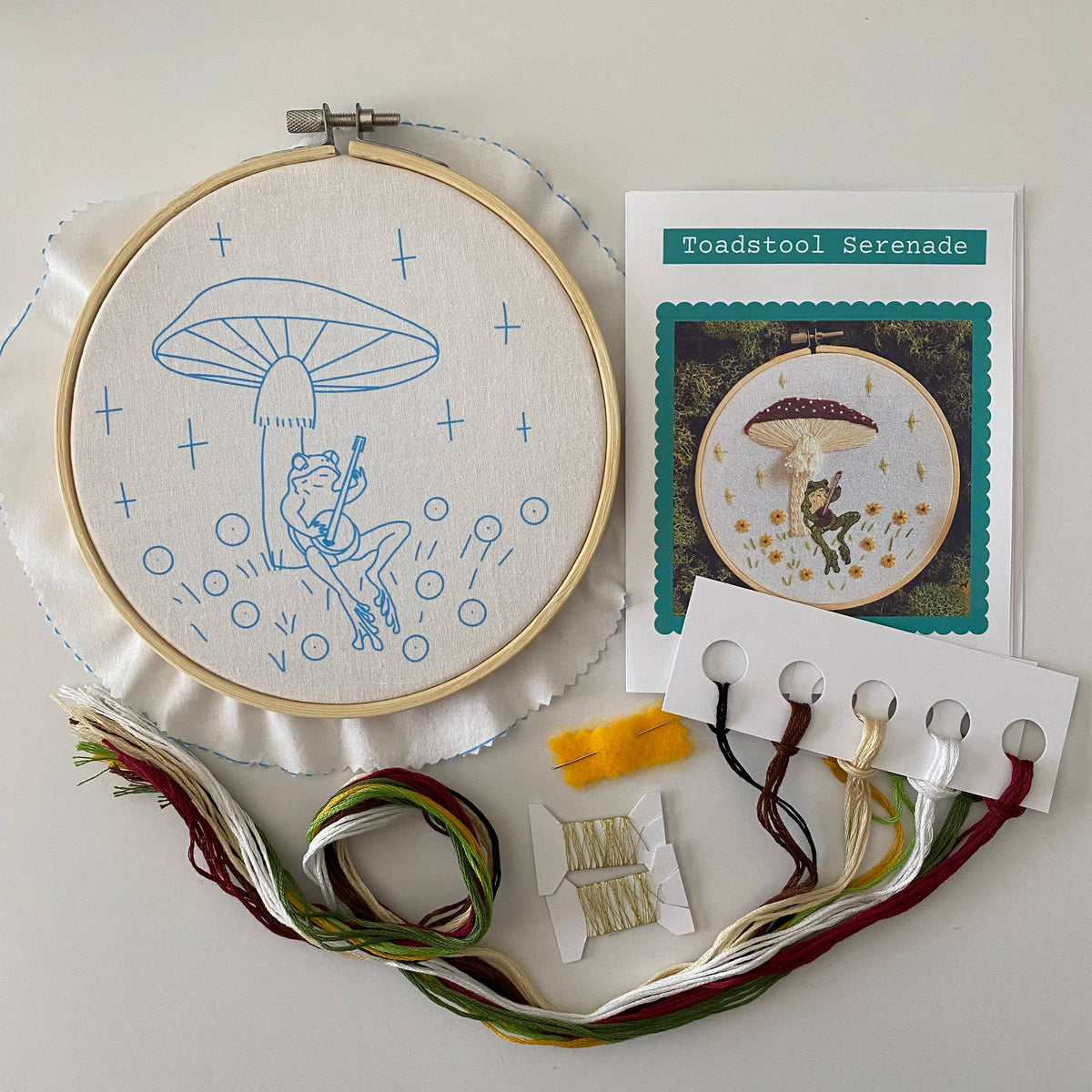 The 6 Beginner Toadstool Serenade Frog & Mushroom DIY Embroidery Kit by Stitches By Tiff features a blue frog on a swing under a mushroom and includes fabric, threads, needle, instructions, and a sample image.