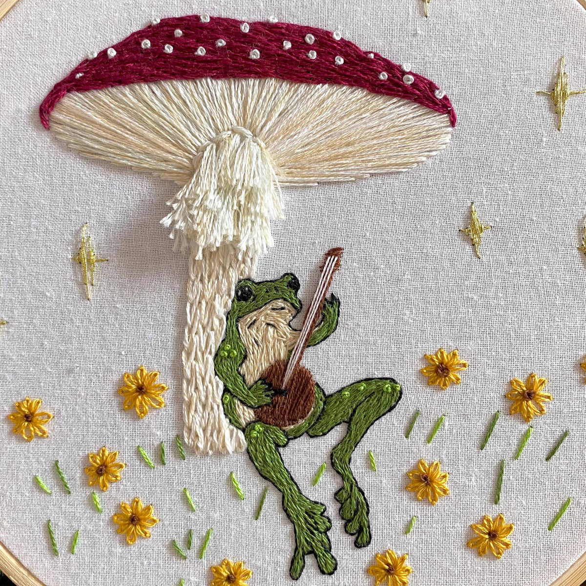 Stitches By Tiff’s 6 Beginner Toadstool Serenade Frog & Mushroom DIY Embroidery Kit features a green frog with guitar under a red-and-white mushroom, surrounded by yellow flowers and stars—perfect for charming DIY art projects.