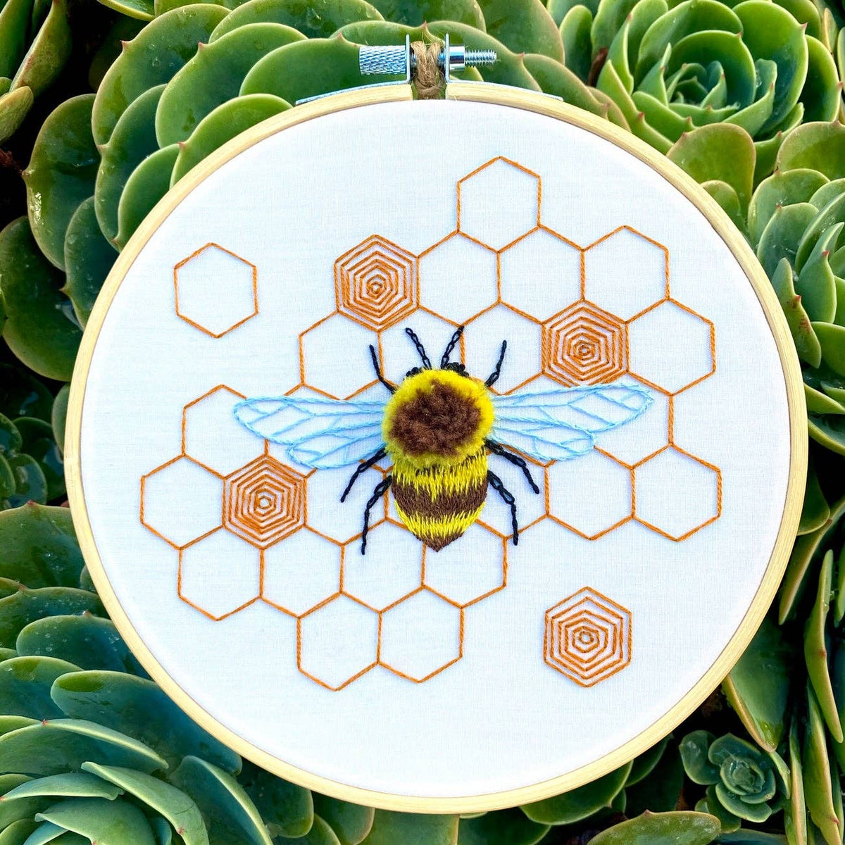 The 6 Fuzzy Bumblebee DIY Embroidery Kit by Stitches By Tiff features a bee with blue wings on white fabric, framed by orange honeycomb in a hoop—ideal for beginners or DIY art projects and displayed among green succulents.