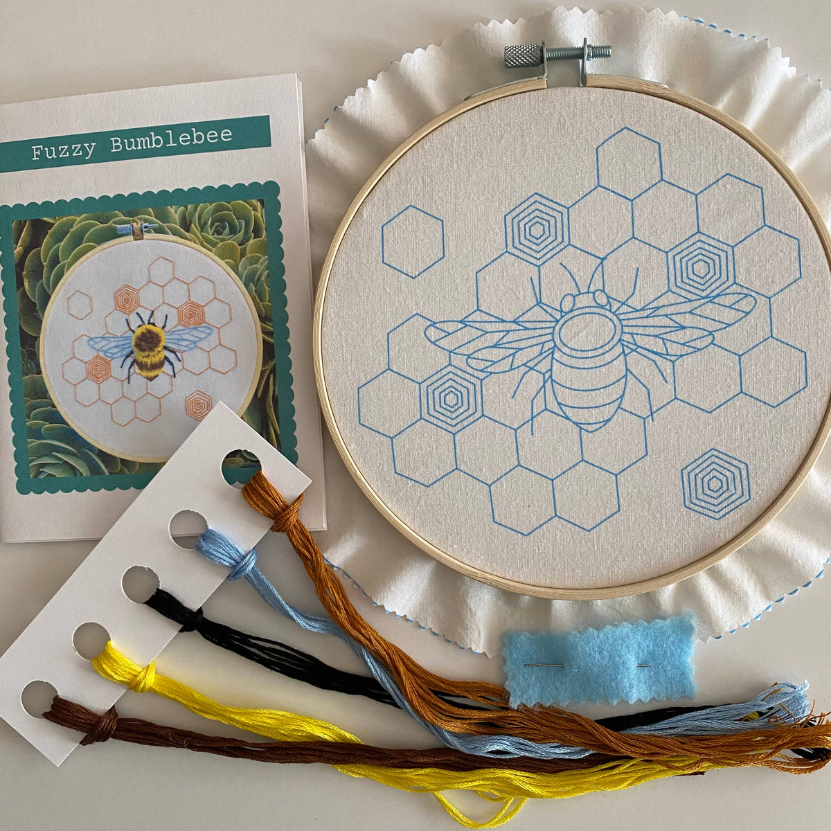 The 6 Fuzzy Bumblebee DIY Embroidery Kit by Stitches By Tiff is ideal for beginners, including a hoop with bee and hexagon pattern, brown, yellow, blue, and black threads, blue felt, and a Fuzzy Bumblebee instruction card.