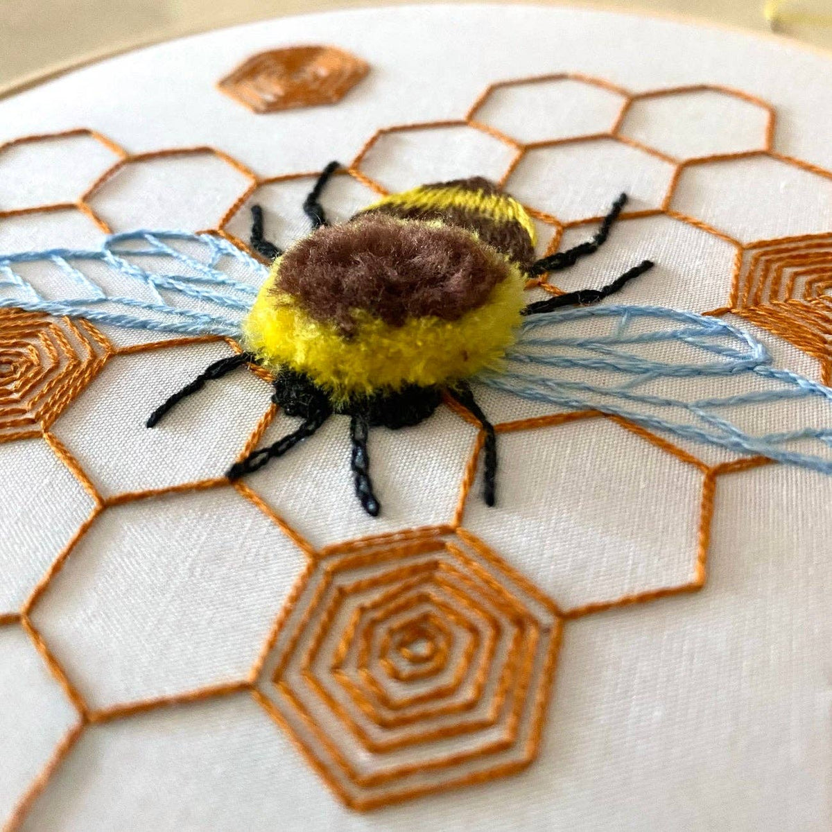 The 6 Fuzzy Bumblebee DIY Embroidery Kit by Stitches By Tiff features a fluffy embroidered bee with blue wings, black legs, and orange honeycomb—perfect for beginner stitchers or creative DIY art projects on white fabric.