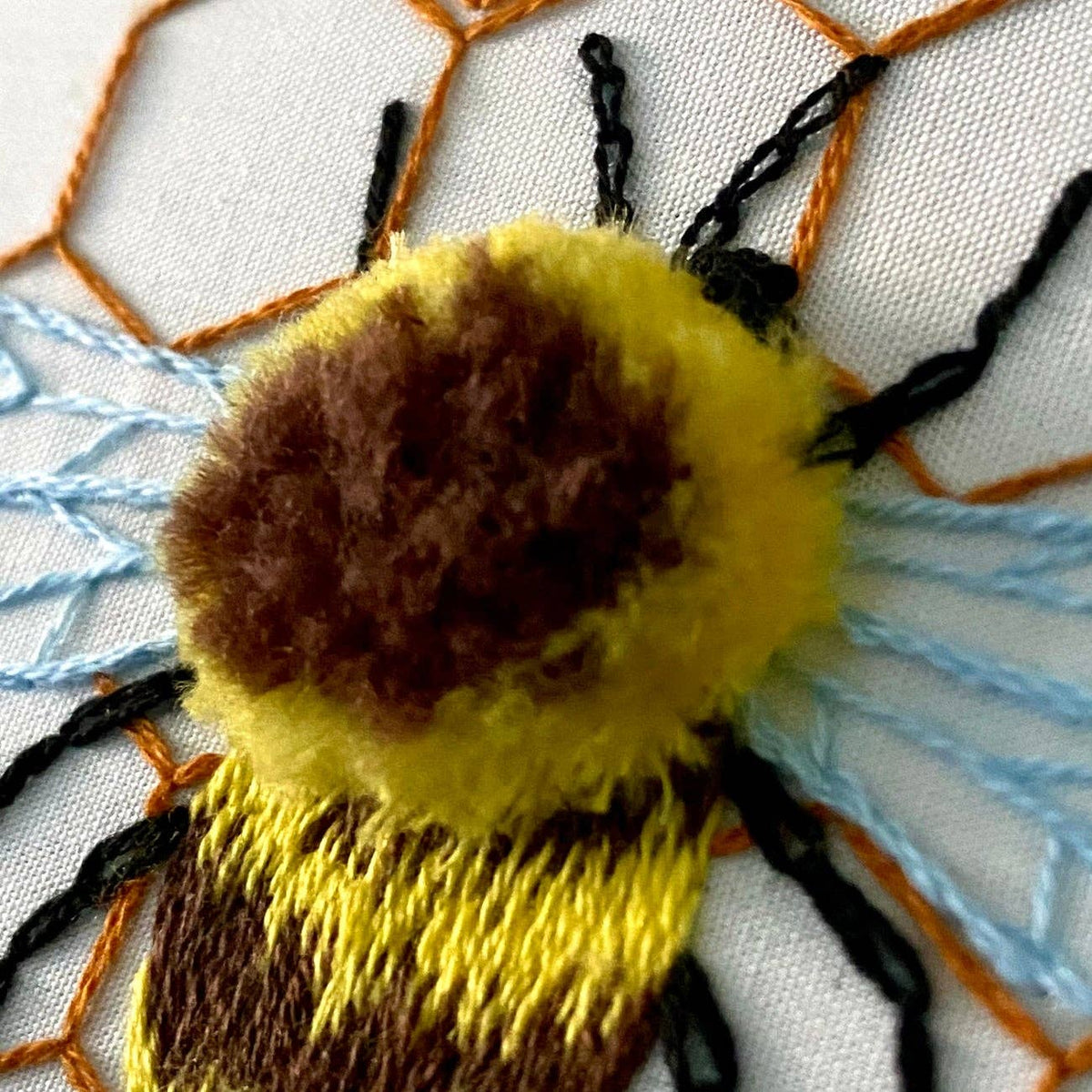 Close-up of the Stitches By Tiff 6 Fuzzy Bumblebee DIY Embroidery Kit: yellow and brown threads form a fuzzy bee on white fabric, with orange and blue stitching for honeycomb and wings—great for beginners or anyone wanting a fun new kit.