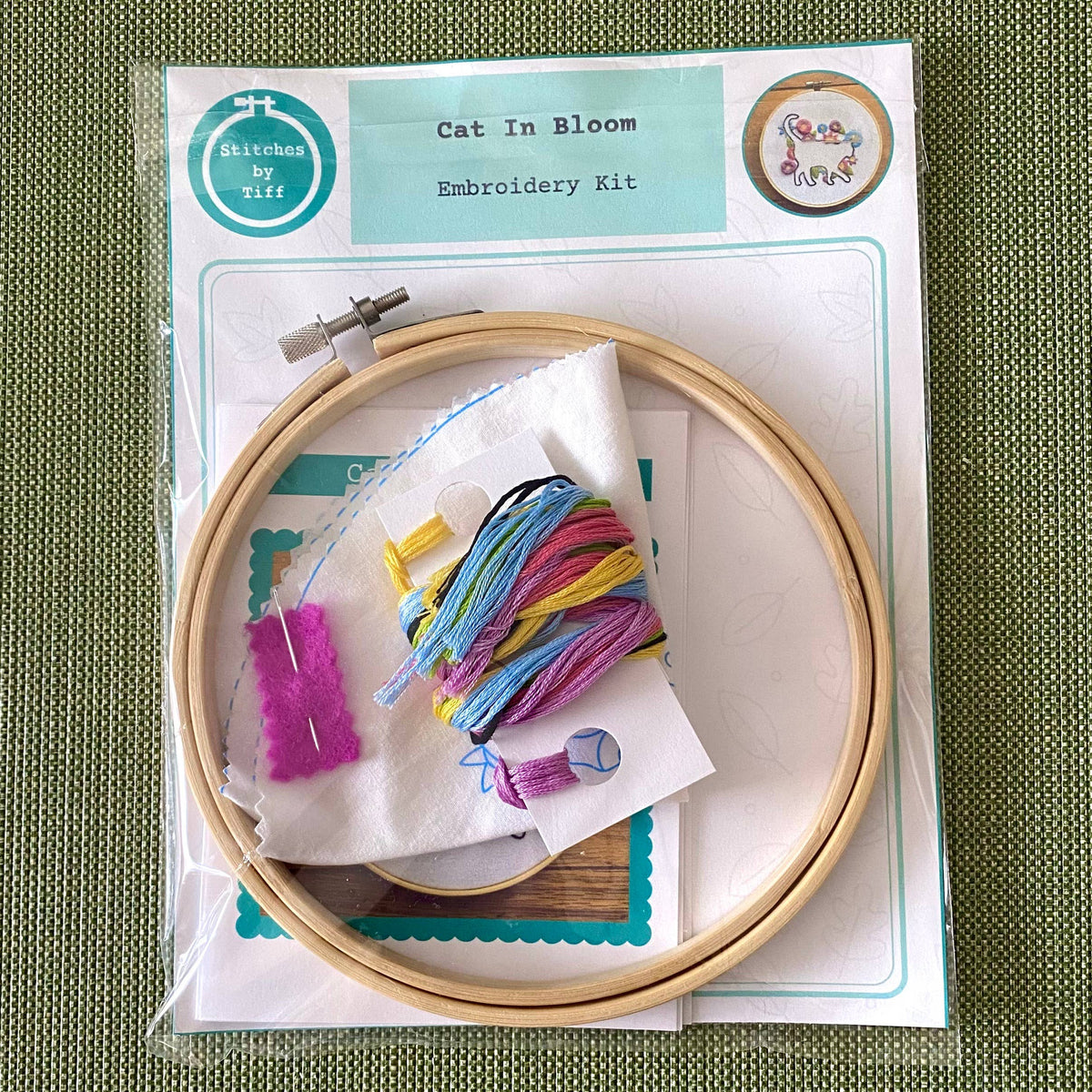 The 6 Beginner Cat in Bloom Silhouette Sampler Embroidery Kit by Stitches By Tiff includes a floral pattern, wooden hoop, colorful threads, needle, fabric, and instructions—ideal for DIY art projects on a green textured surface.