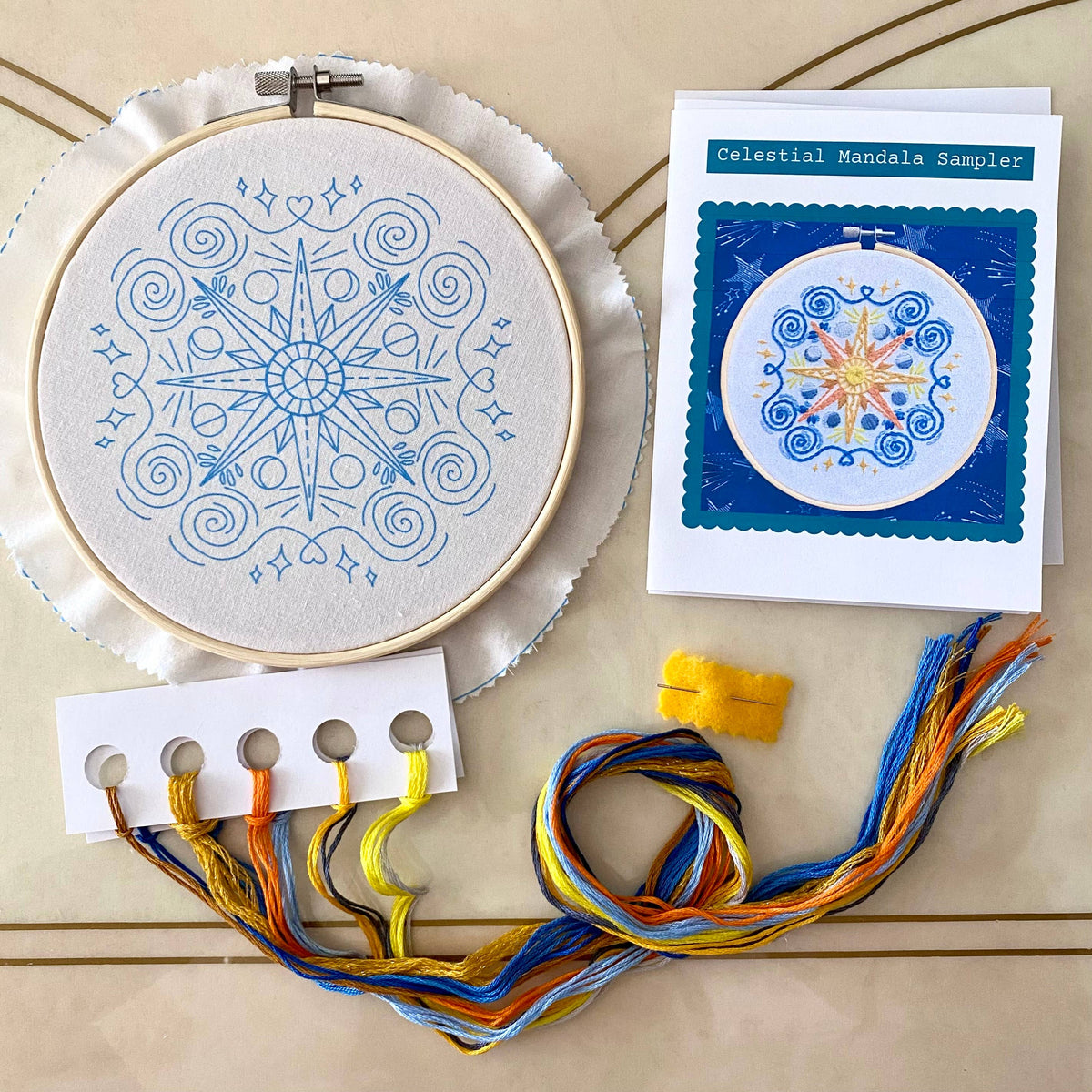 The Stitches By Tiff 6 Beginner Celestial Mandala Sampler Embroidery Kit includes a hooped fabric with a blue mandala pattern, sample card, colored floss, needle, and threader—ideal for beginner embroidery and DIY art projects.