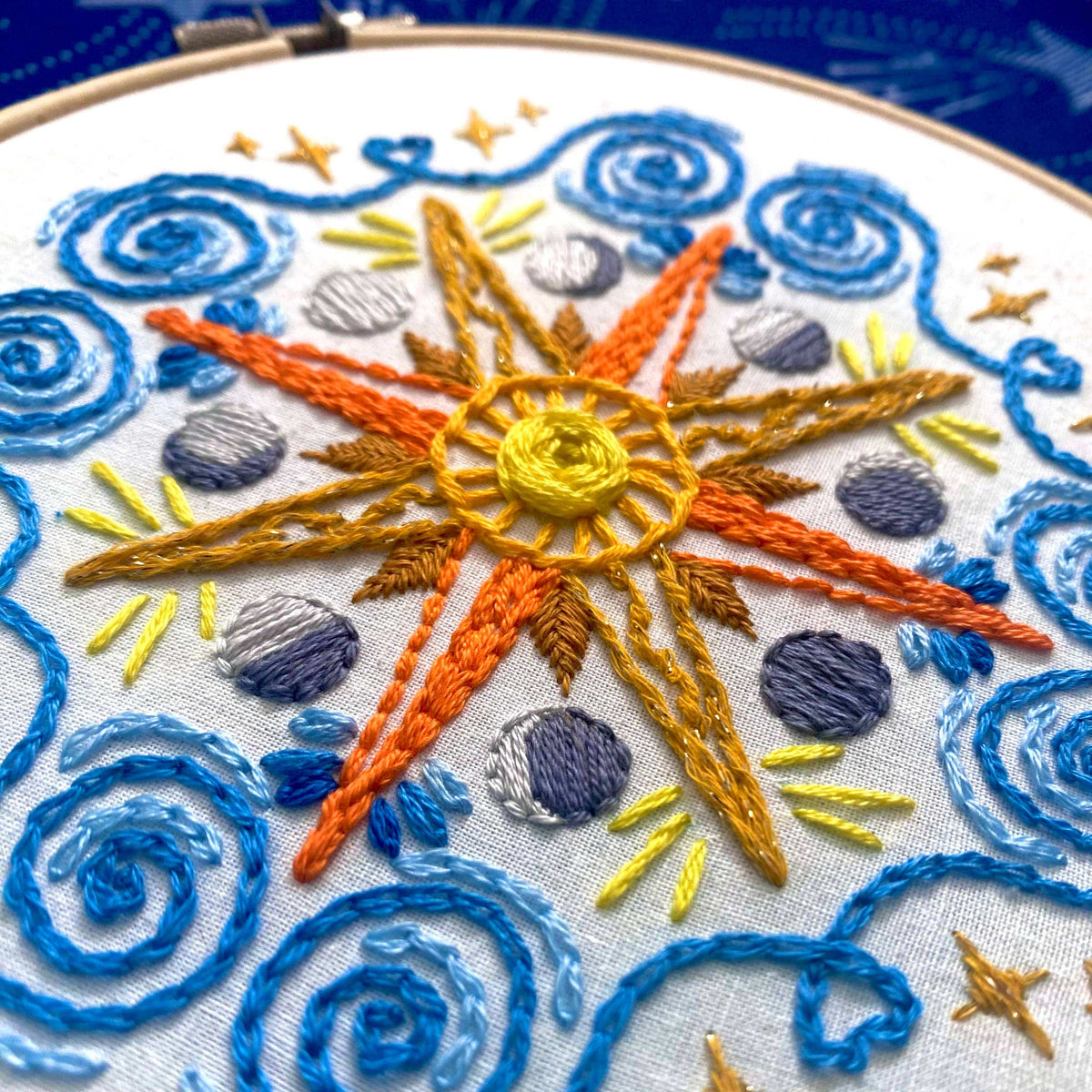 The Stitches By Tiff 6 Beginner Celestial Mandala Sampler Embroidery Kit features colorful sun embroidery with orange and yellow rays, blue swirls, gray circles, and star details on white fabric—ideal for beginner DIY art projects.