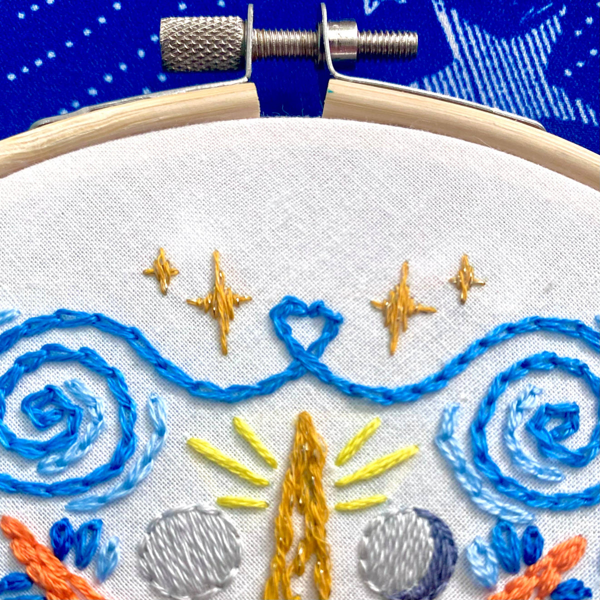 Close-up of the Stitches By Tiff 6 Beginner Celestial Mandala Sampler Embroidery Kit, showing blue swirls, yellow and gold stars, and gray shapes on white fabric—great for beginner embroidery or DIY art projects. Blue patterned background visible.