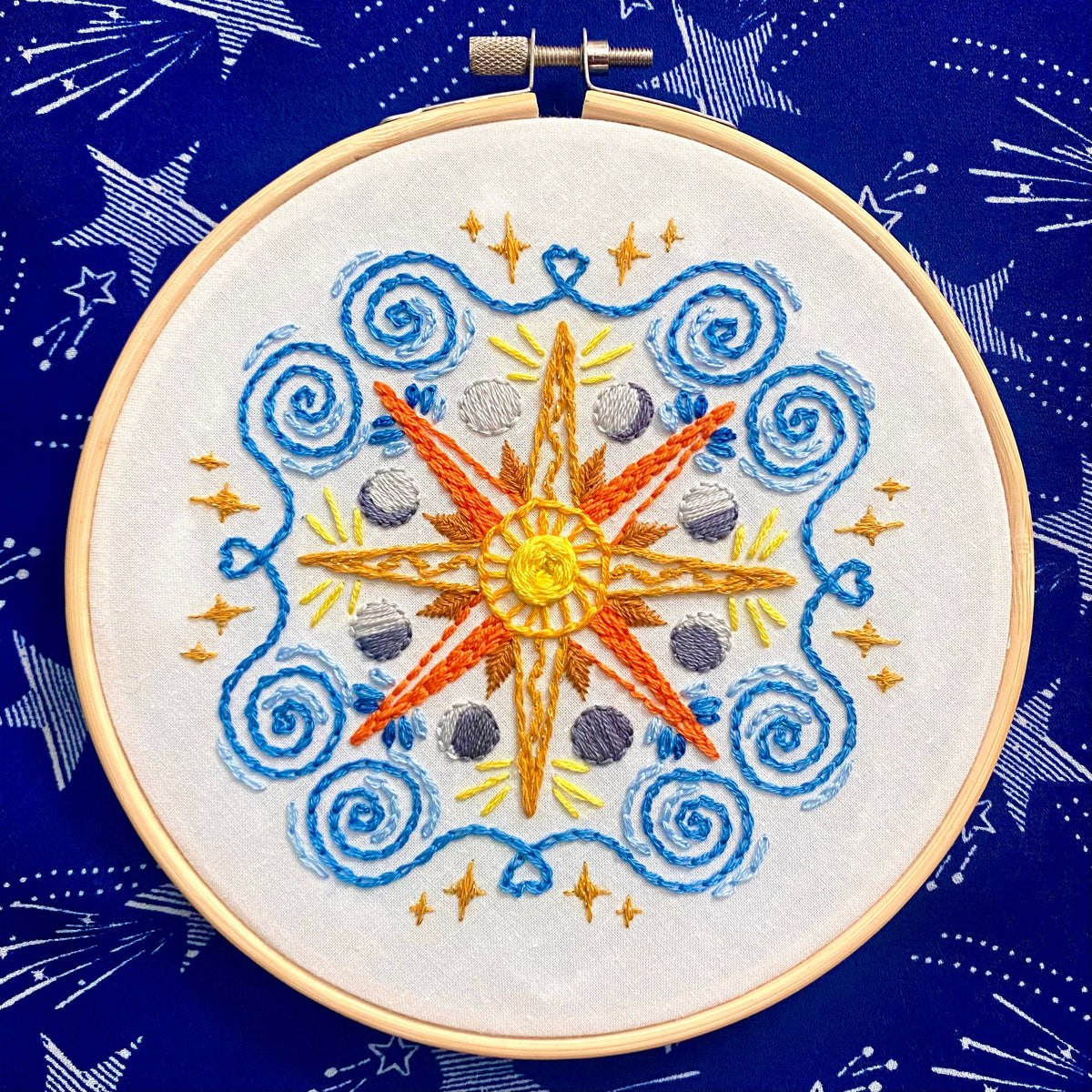 The 6 Beginner Celestial Mandala Sampler Embroidery Kit by Stitches By Tiff features a sun and star center with orange, blue, and yellow celestial patterns on blue fabric—ideal for beginner embroidery or DIY art projects.