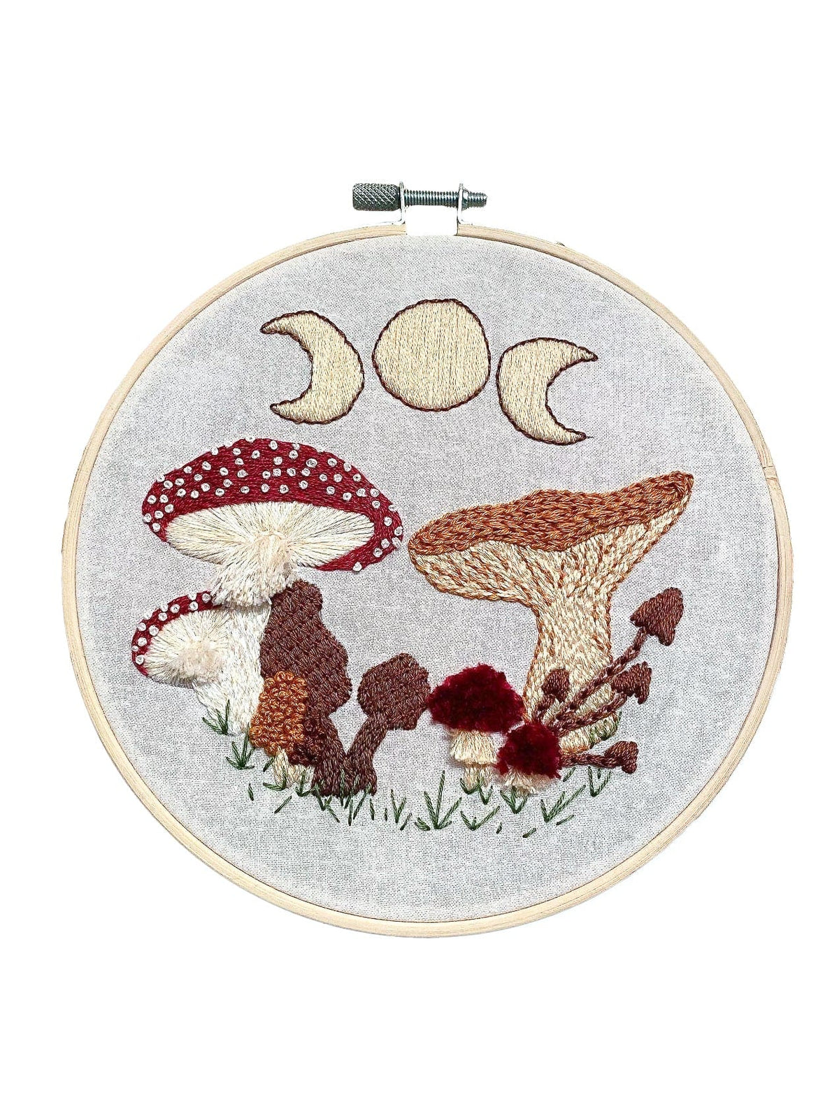 The 6 Beginner Mushie DIY Embroidery Kit by Stitches By Tiff features a brown and red mushroom with green grass and moon phases on light fabric—perfect for adding a magical touch to your space or starting embroidery projects.