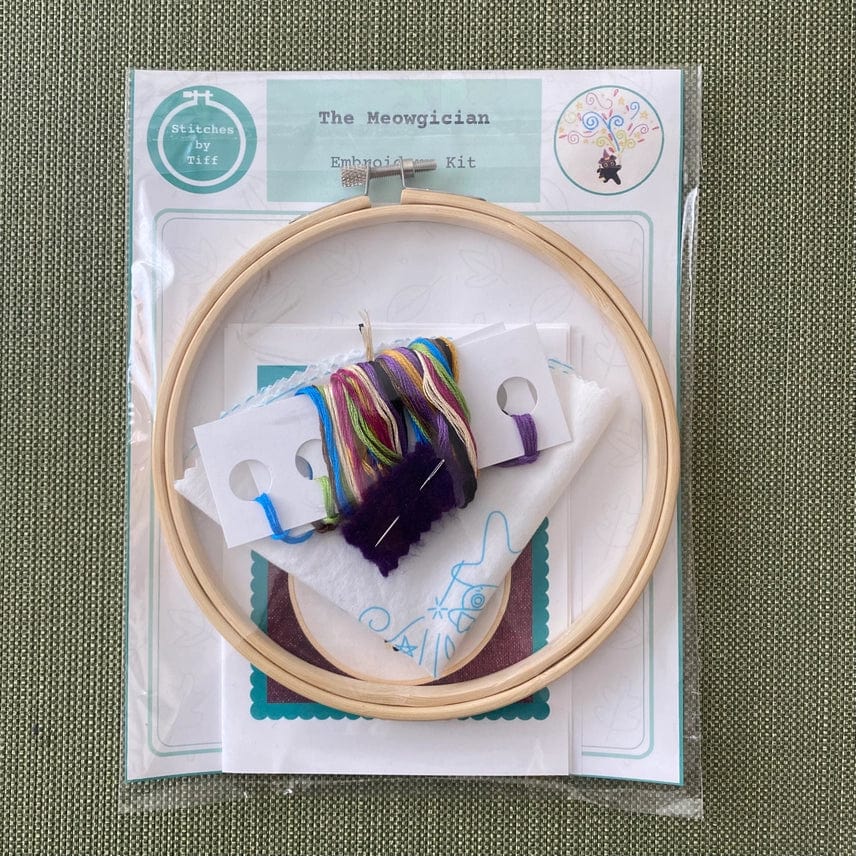 Stitches By Tiff’s 6 Beginner The Meowgician Embroidery Kit includes a black cat pattern, wooden hoop, multicolored thread, folded fabric, needle, and instructional card—neatly packaged on a green textured surface.