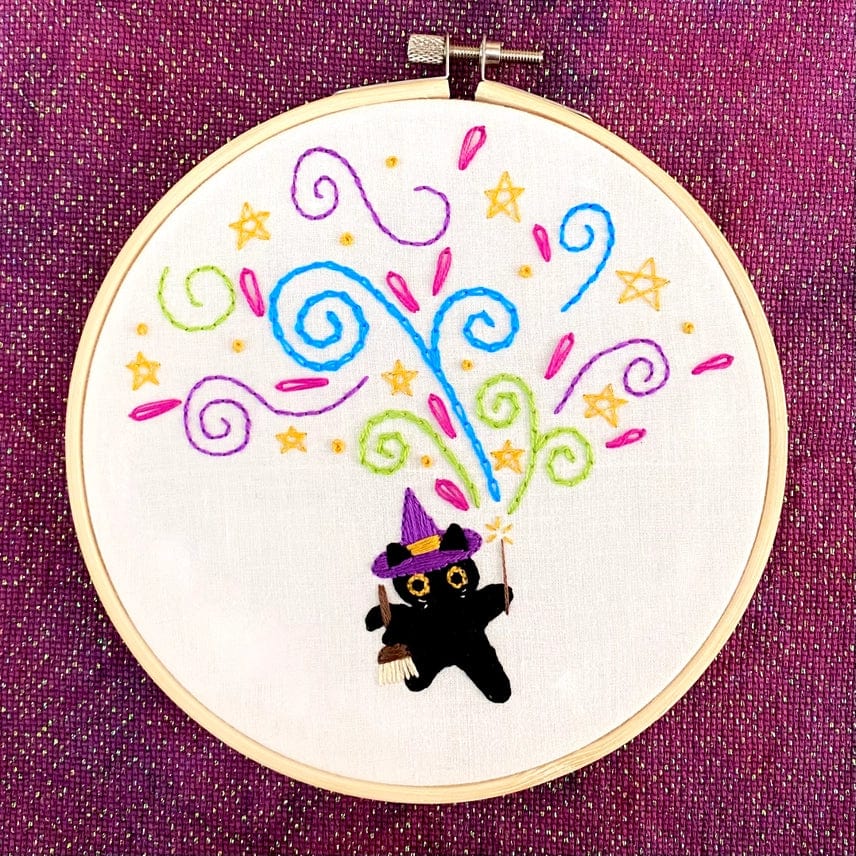 The 6 Beginner The Meowgician Embroidery Kit by Stitches By Tiff features a cute black cat in a witch hat with a wand, colorful swirls, stars, and sparkles on white fabric in a hoop—perfect for beginners.