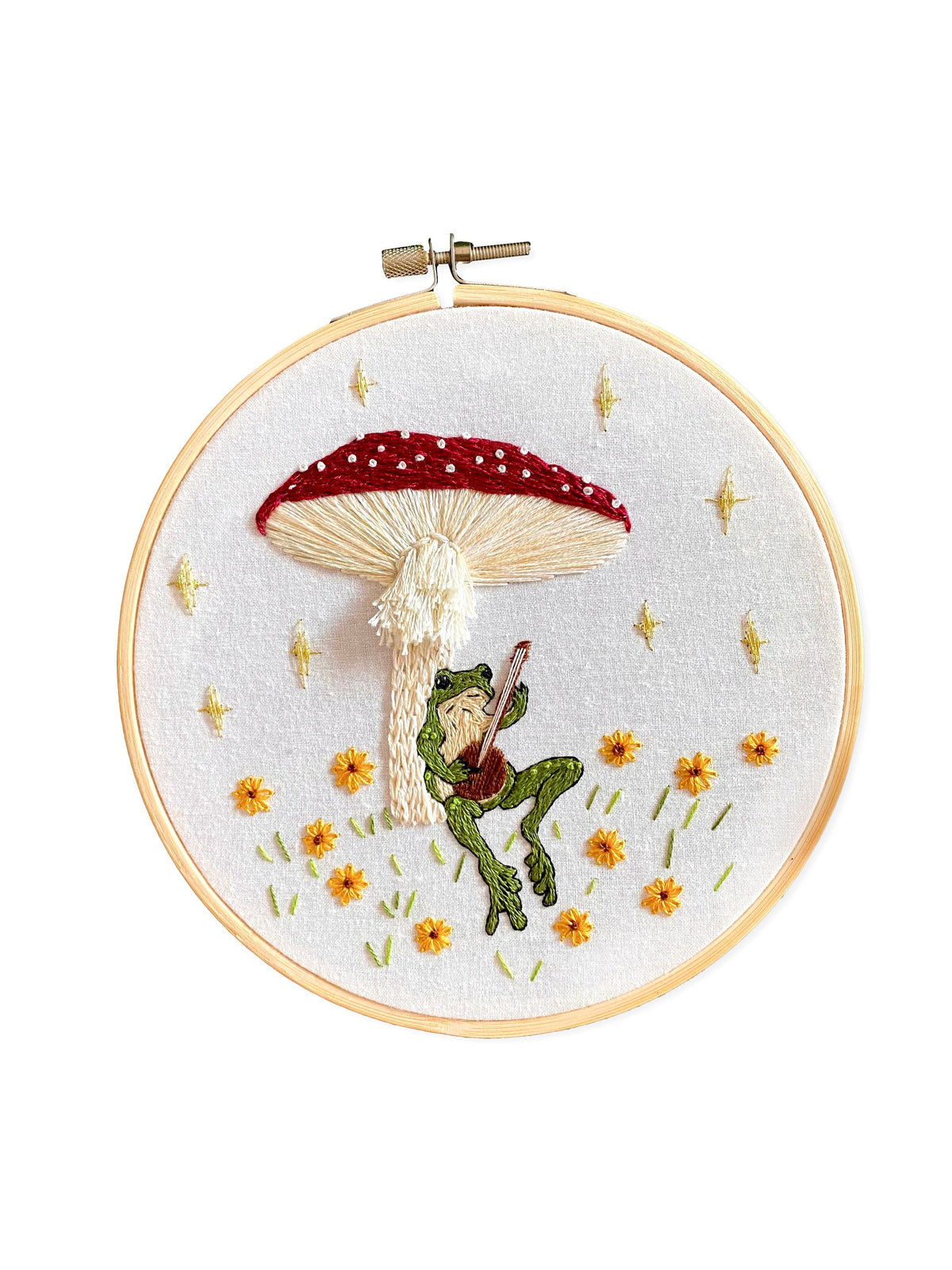The 6 Beginner Toadstool Serenade Frog & Mushroom DIY Embroidery Kit by Stitches By Tiff features a guitar-playing frog under a red-and-white mushroom, with yellow flowers and stars—a delightful piece for DIY art projects.