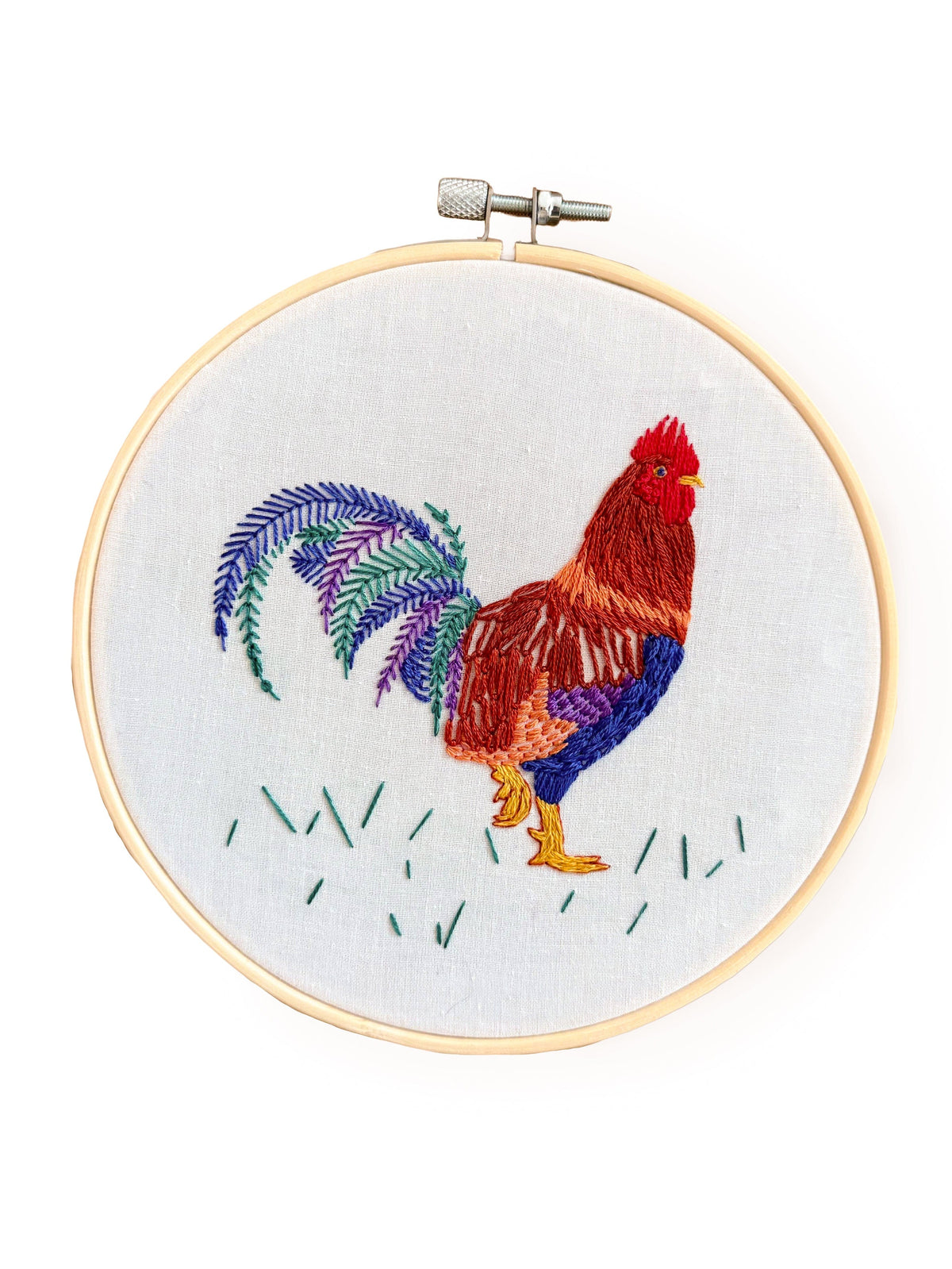 The Stitches By Tiff 6 Rustic Rooster Embroidery Kit features a vibrant rooster on white fabric, accented with green grass and colorful tail feathers—an eye-catching DIY art project for your home.