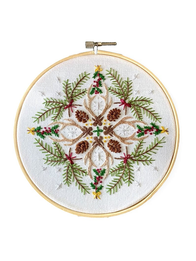 The Stitches By Tiff 6 Winter Woodland Mandala DIY Embroidery Kit features a symmetrical winter design with pinecones, green pine branches, red berries, and yellow stars on white fabric—ideal for creative art projects or as a Woodland Mandala Sampler.