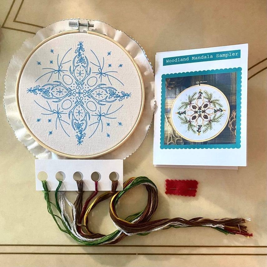 The Stitches By Tiff 6 Winter Woodland Mandala DIY Embroidery Kit includes fabric with a blue mandala, colorful threads, a thread holder, red felt piece, and an instruction booklet—ideal for creative DIY art projects.