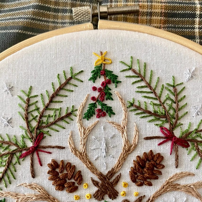 The Stitches By Tiff 6 Winter Woodland Mandala DIY Embroidery Kit features pine branches, cones, red berries, green leaves, and brown antlers on cream fabric—ideal for embroidery enthusiasts. A plaid cloth background adds a cozy touch.