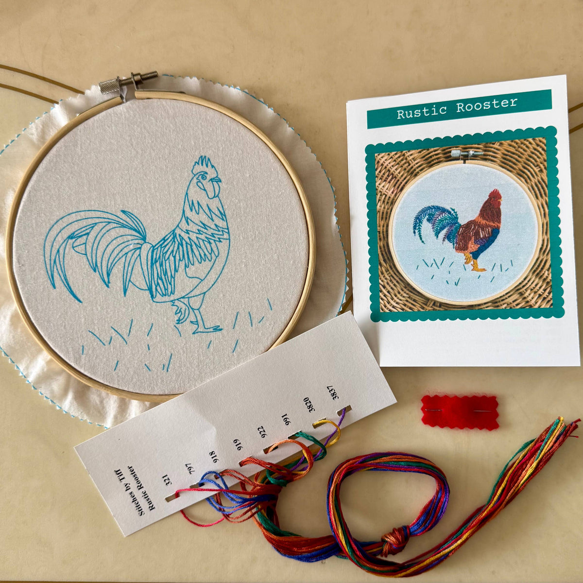 A rooster embroidery project from the Stitches By Tiff 6 Rustic Rooster Embroidery Kit is shown in progress with a hoop, colorful threads, partially stitched fabric, and an instruction card displaying the completed design.