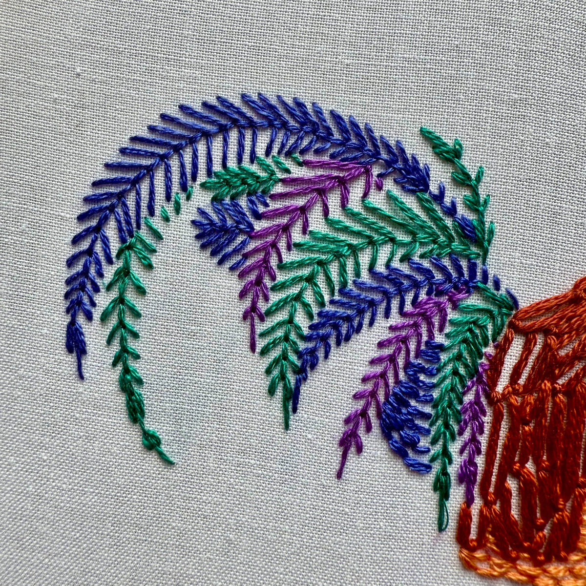 Colorful fern-like leaves in green, purple, and blue thread on white fabric with an orange-brown accent at the corner—ideal inspiration for DIY art or the 6 Rustic Rooster Embroidery Kit from Stitches By Tiff.