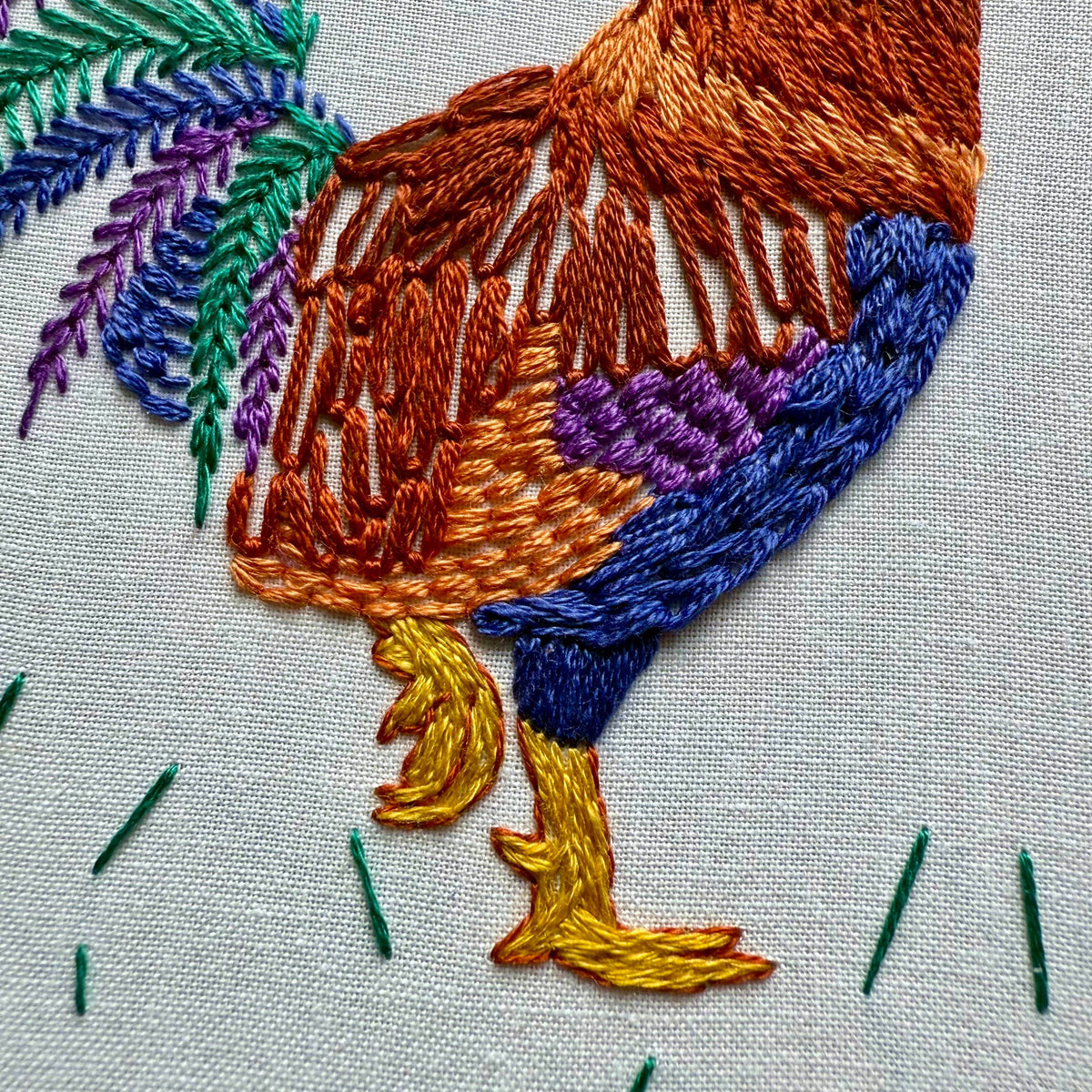 Close-up of the Stitches By Tiff 6 Rustic Rooster Embroidery Kit, featuring vibrant yellow legs, blue and purple body, orange and brown feathers, and a green tail on light fabric—ideal for creative DIY art projects.