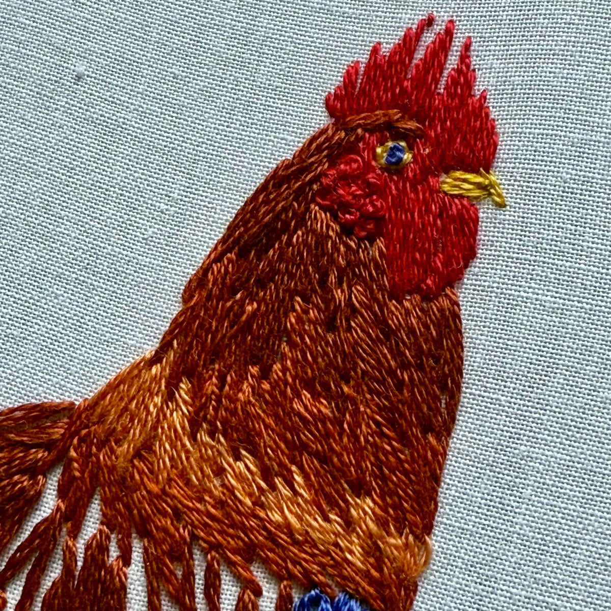 The Stitches By Tiff 6 Rustic Rooster Embroidery Kit features an embroidered rooster with a red comb, brown and orange feathers, and a yellow beak on light fabric—perfect for DIY art projects or rustic decor lovers.