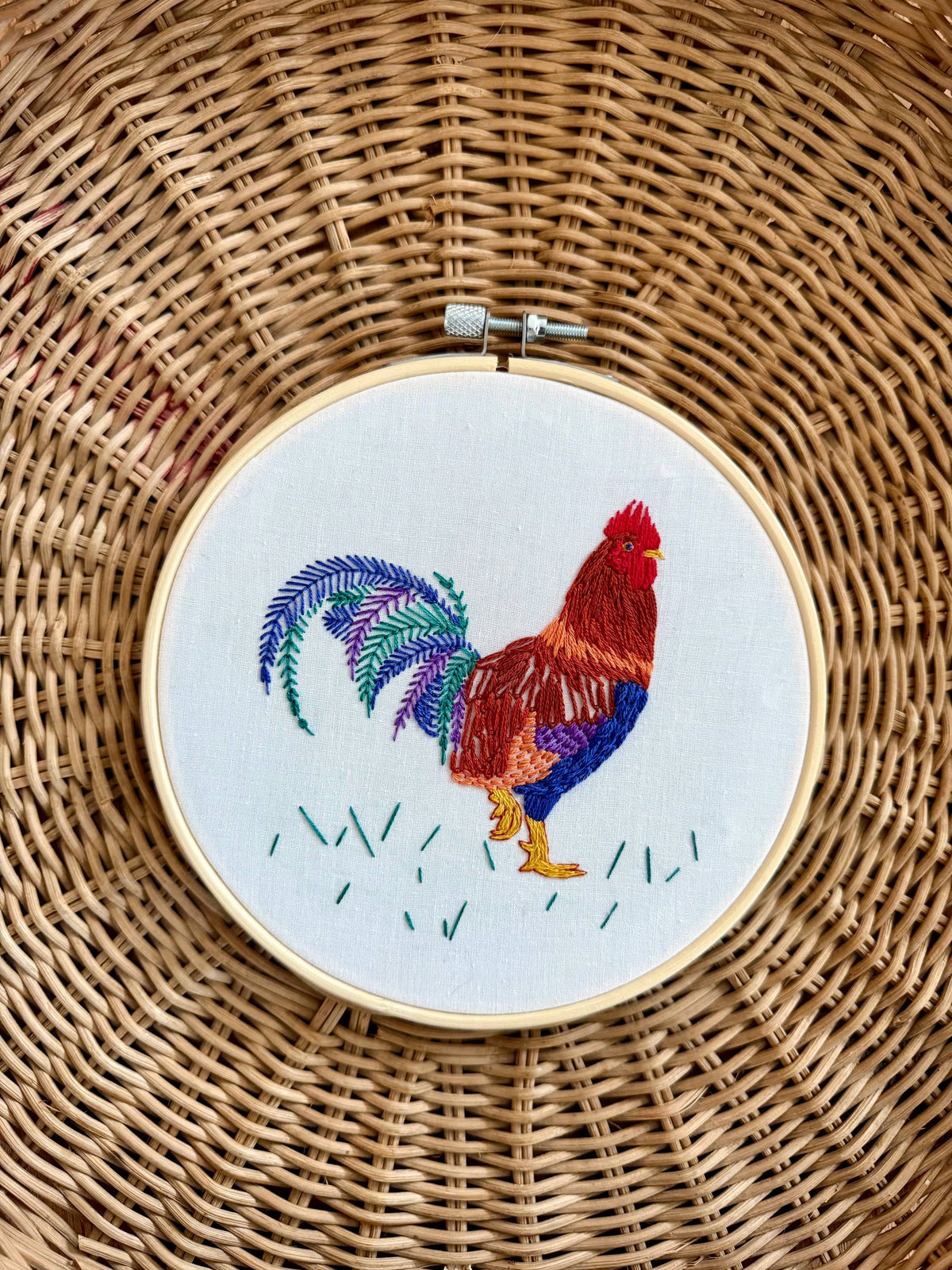 The 6 Rustic Rooster Embroidery Kit by Stitches By Tiff features a colorful, detailed rooster on white fabric with stitched grass, perfect for DIY art projects. Displayed on a woven wicker surface for added texture.
