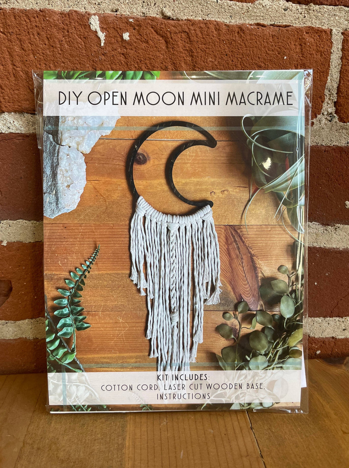 The DIY Open Moon Beginner Mini Macrame Kit by Tangled Up In Hue features a crescent moon wooden base, white cords, and instructions—ideal for crafting a unique wall hanging. Displayed with plants and brick wall. Text: DIY OPEN MOON MINI MACRAME.