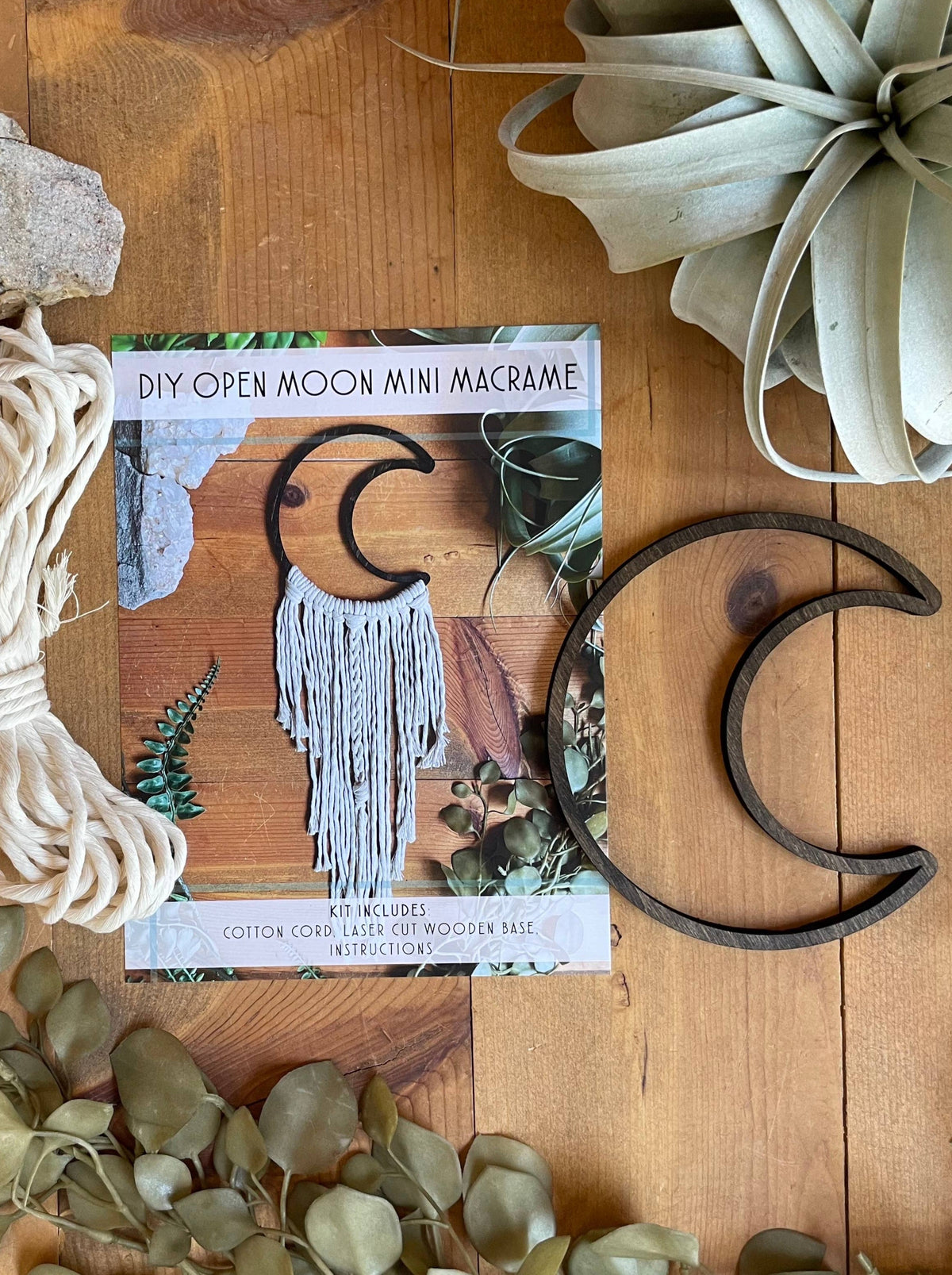 The DIY Open Moon Beginner Mini Macrame Kit by Tangled Up In Hue includes a wooden crescent moon base, white cotton cord, and an instruction card with a finished sample—perfect for wall hanging projects.