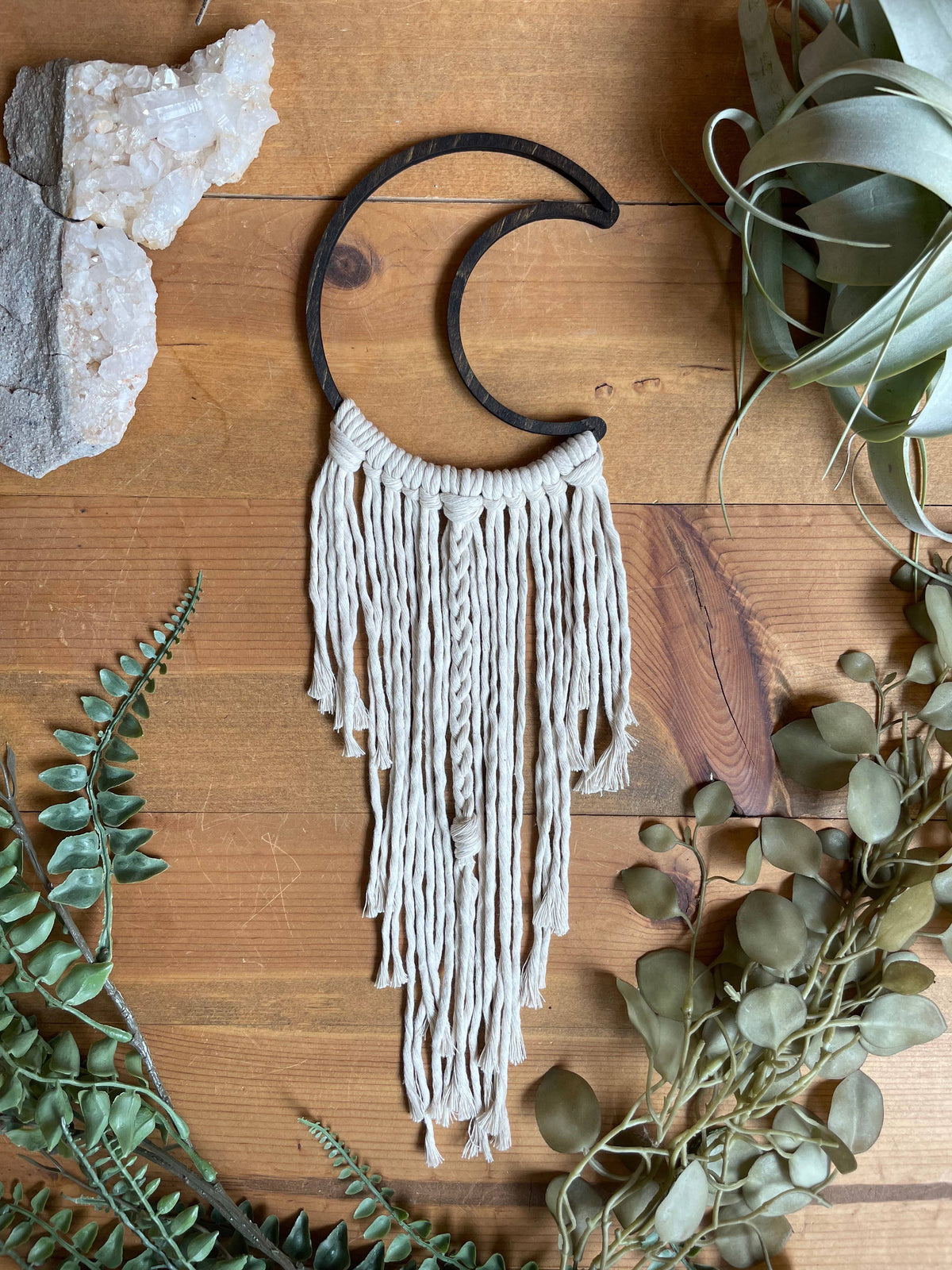 The DIY Open Moon Beginner Mini Macrame Kit by Tangled Up In Hue features a cream macramé crescent moon, displayed on wood amid greenery, air plants, and white crystals—perfect for your next macrame project.