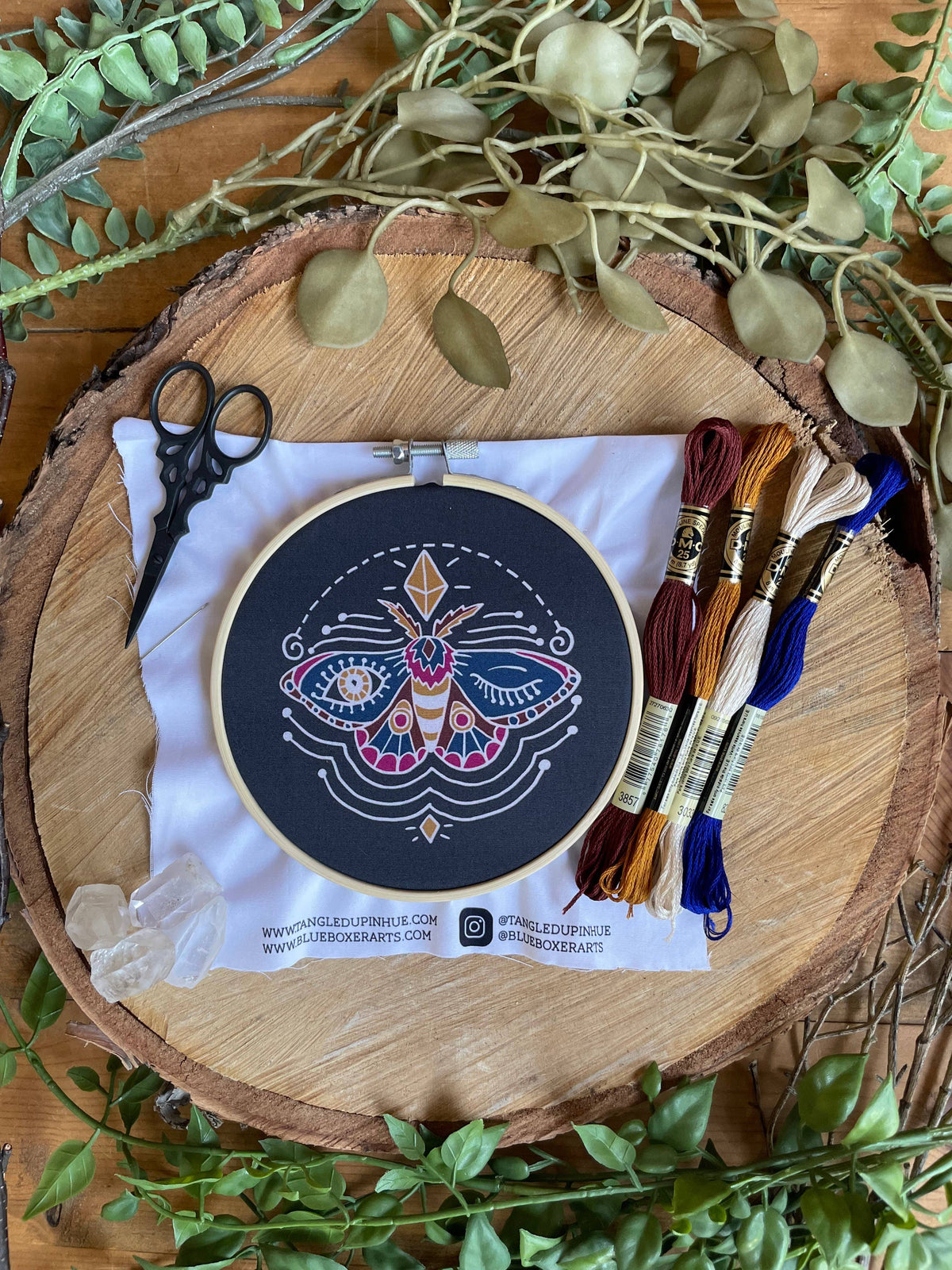 The 5 Beginner Winking Moth Embroidery Kit by Tangled Up In Hue sits on a cloth with thread, scissors, crystals, and leaves—ideal for beginner crafters seeking a colorful project.