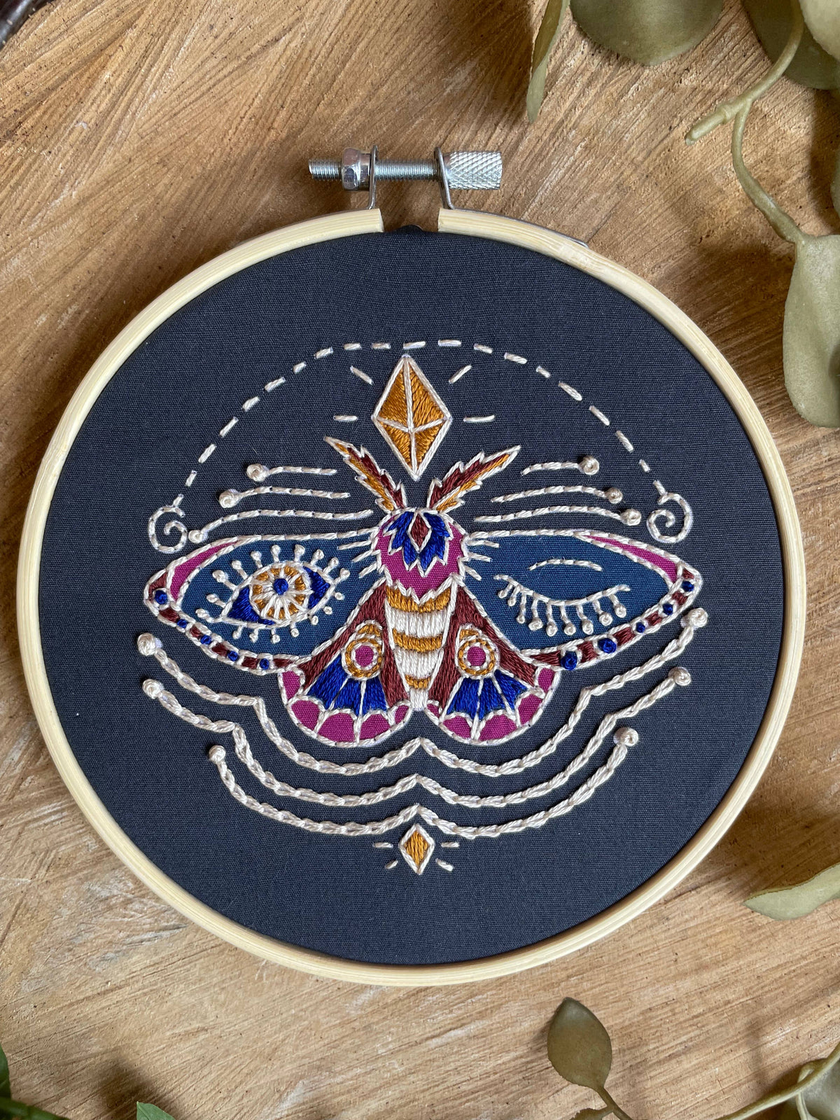 The Tangled Up In Hue 5 Beginner Winking Moth Embroidery Kit features a vibrant moth stitched in gold, blue, purple, and red on black fabric with geometric and celestial accents—ideal for beginner crafters.