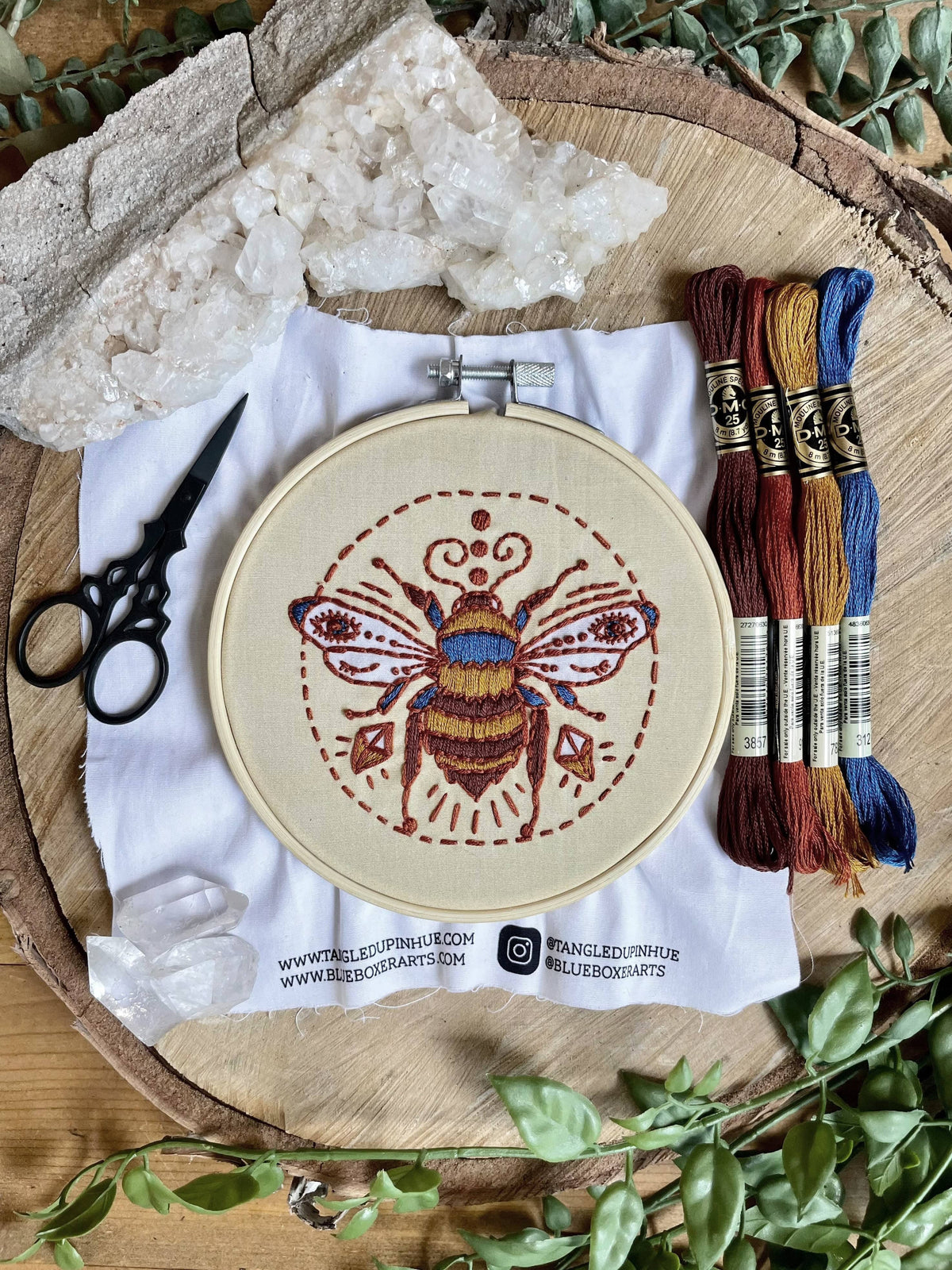 The Tangled Up In Hue 5 Beginner Winking Bee Embroidery Kit includes a bee design in a wooden hoop, earthy-toned floss, black scissors, and crystals—an ideal starter set for new crafters.